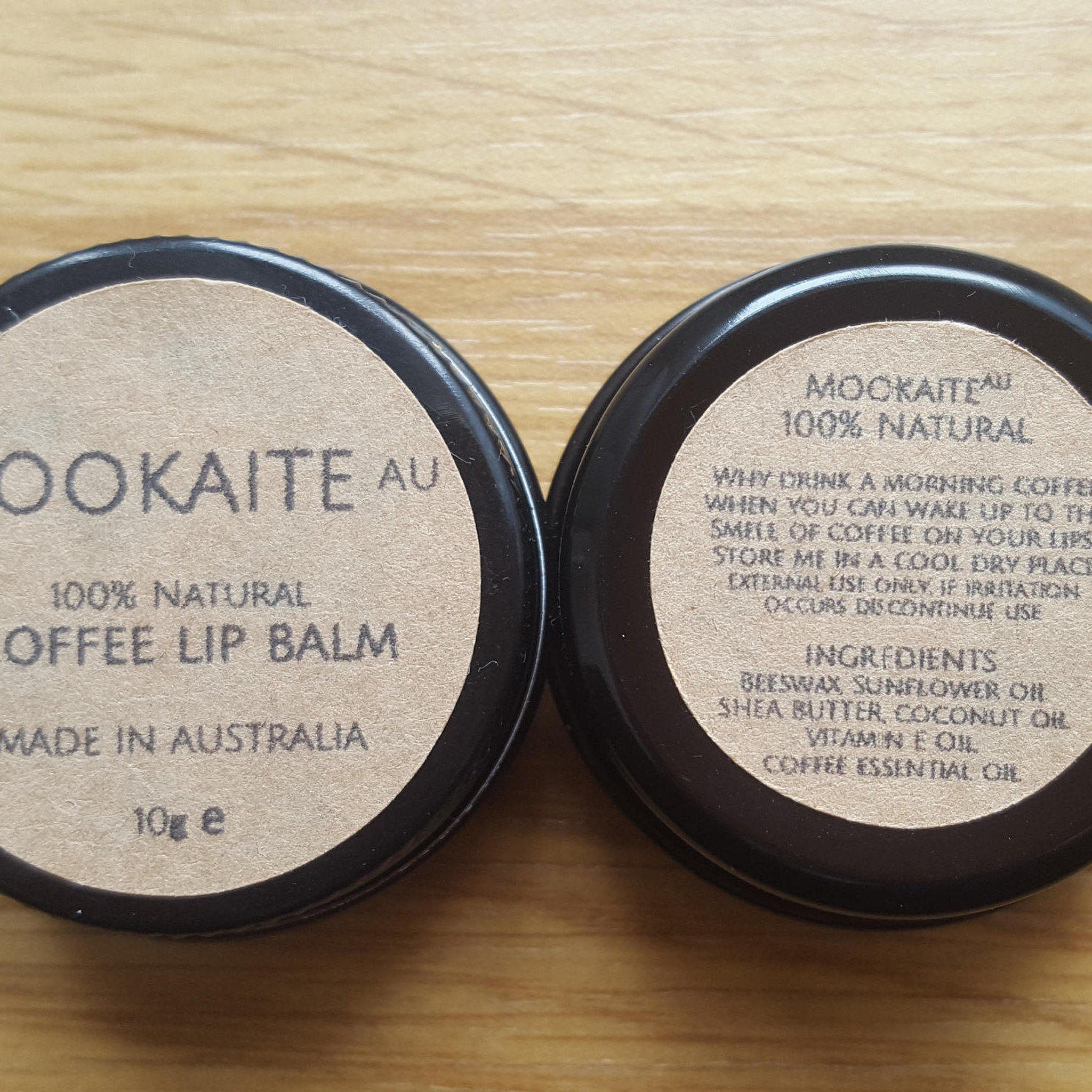 Lip balm tin with black rim, tan label, and visible "Mookaite AU" text on wooden surface