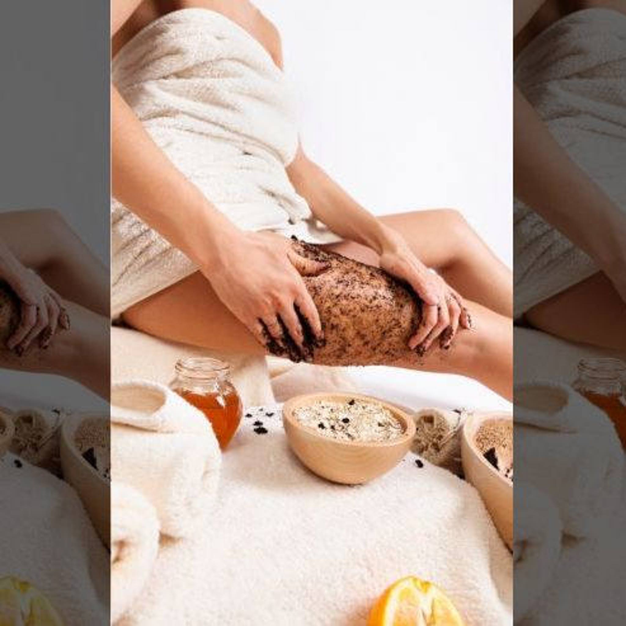 Body scrub application on leg with brown scrub, white towel, and wooden bowl with ingredients