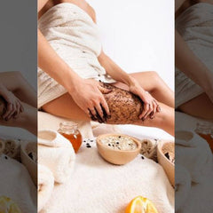 Body scrub application on leg with brown scrub, white towel, and wooden bowl with ingredients