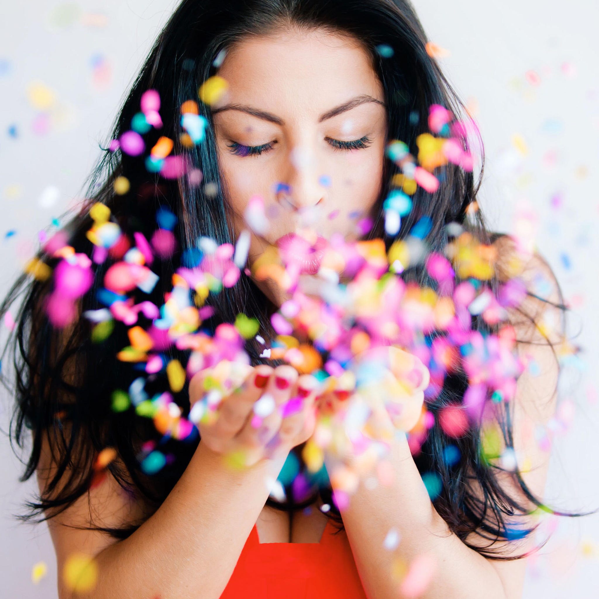 Woman blowing colorful confetti, with black hair and red top, celebratory style