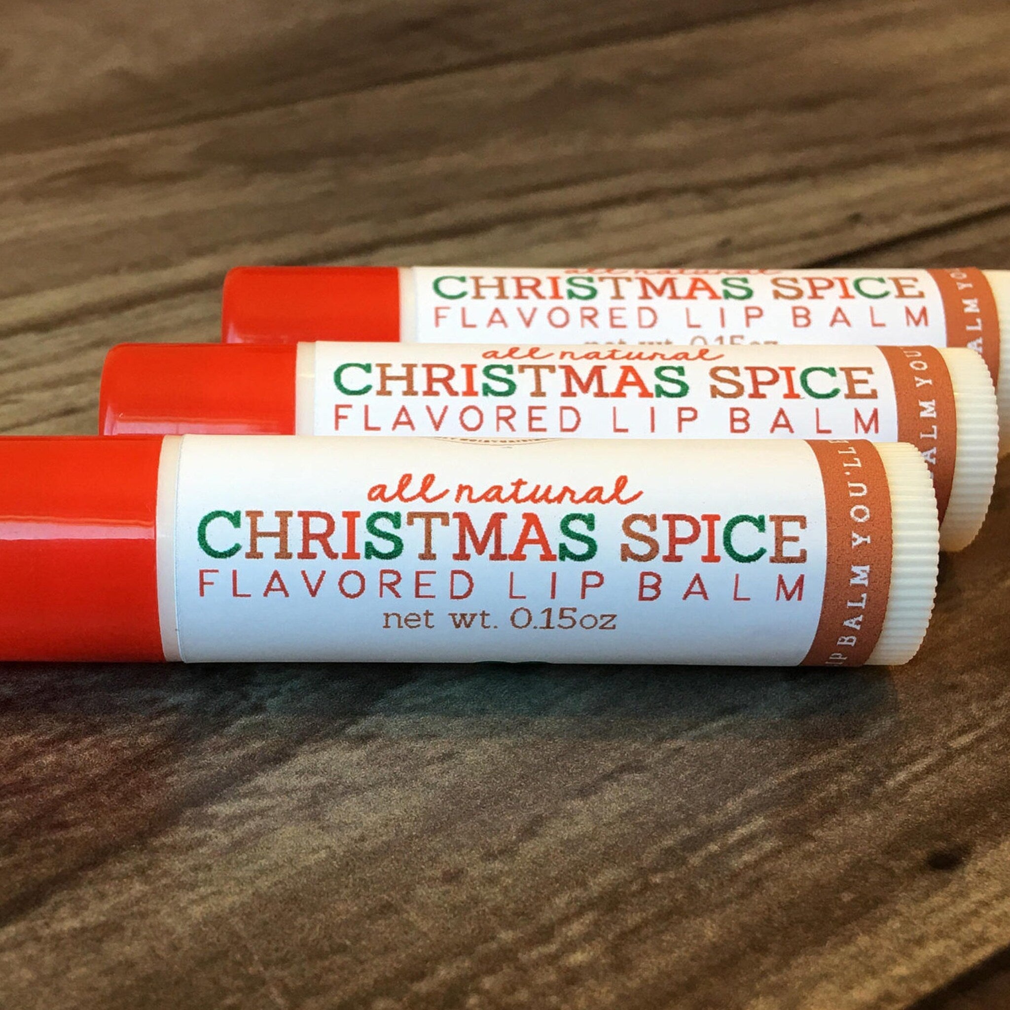 Lip balm tubes with red, white, and brown text, Christmas Spice flavor, on wooden surface