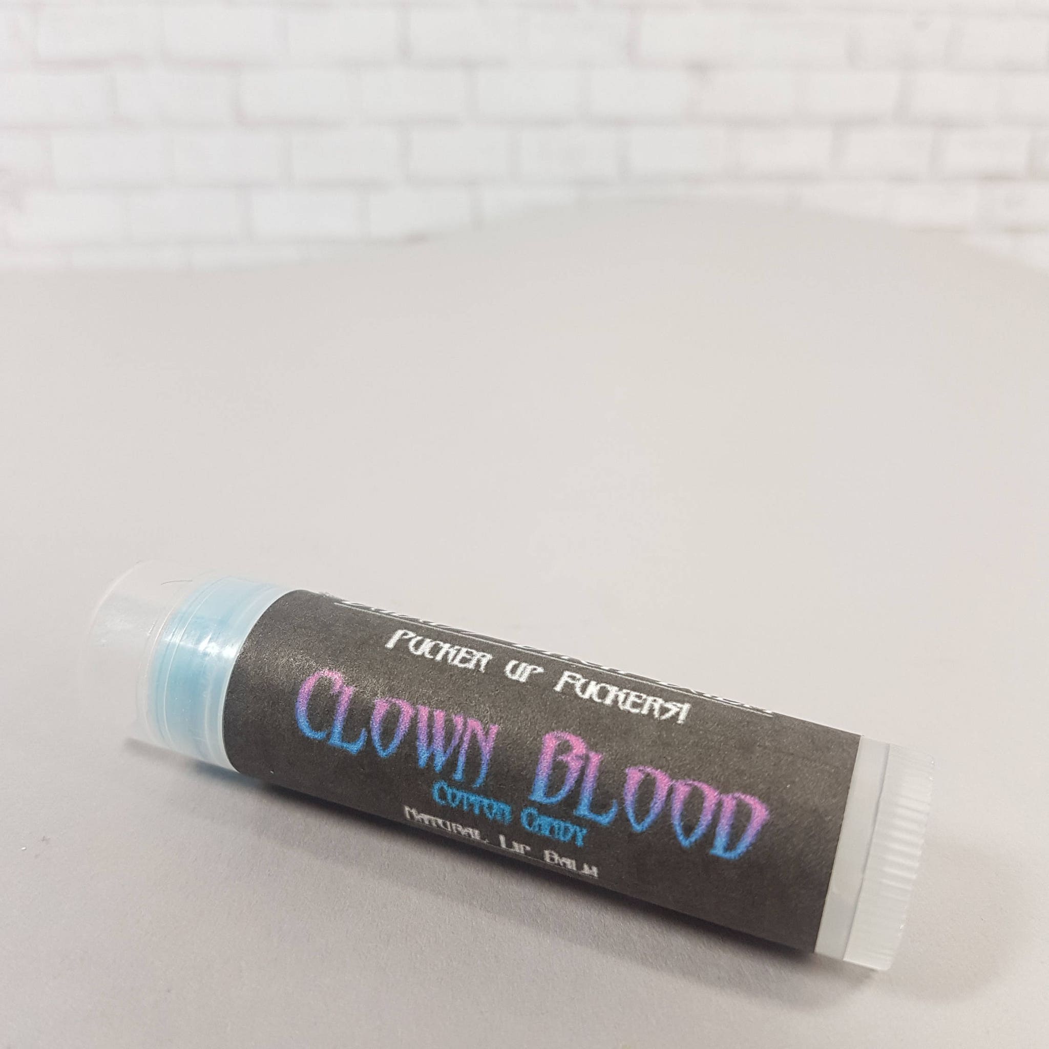 Lip balm tube with black label, blue and pink text, and clear cap, "CLOWN BLOOD" visible