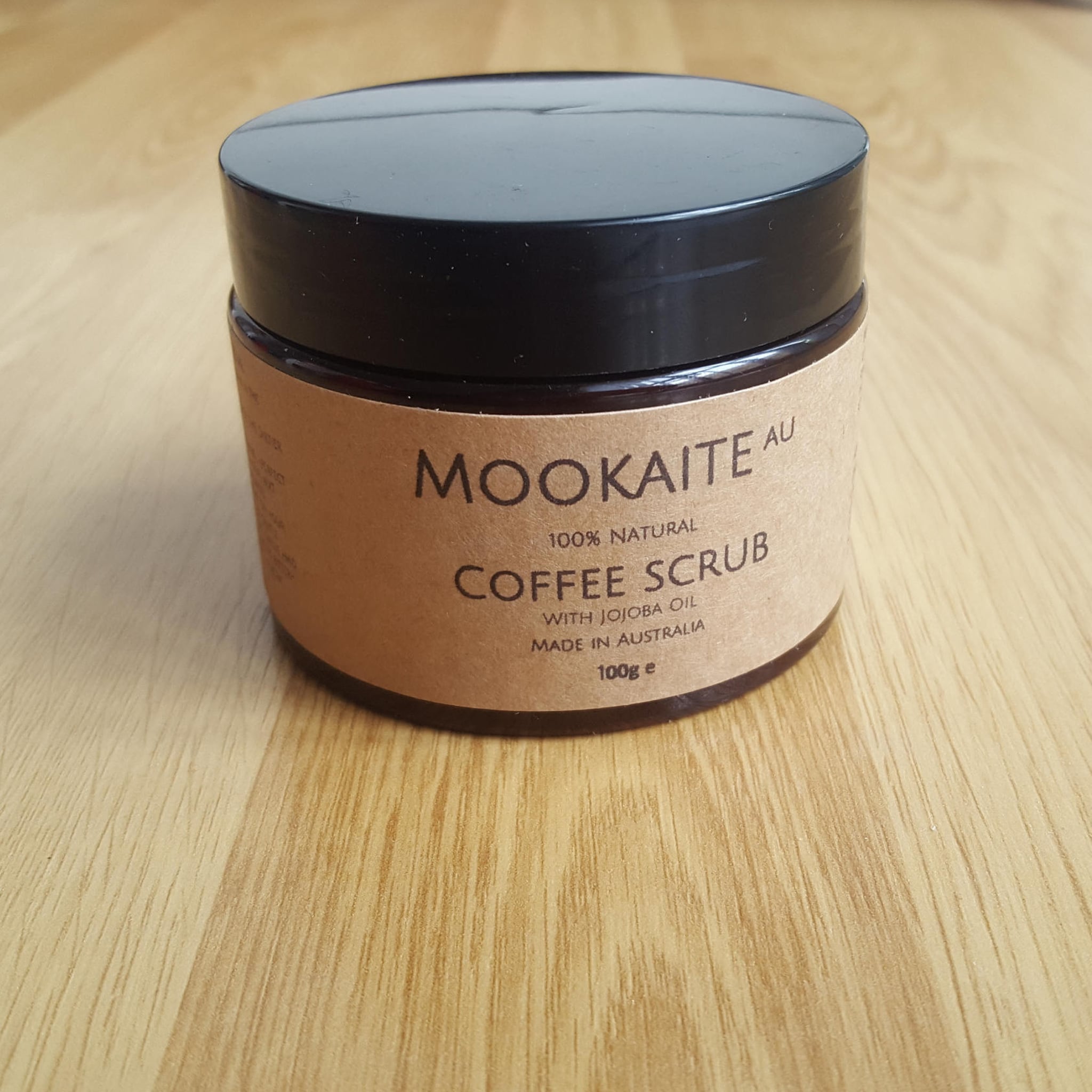 Coffee scrub jar with black lid, brown label, and wooden surface below MOOKAITE brand