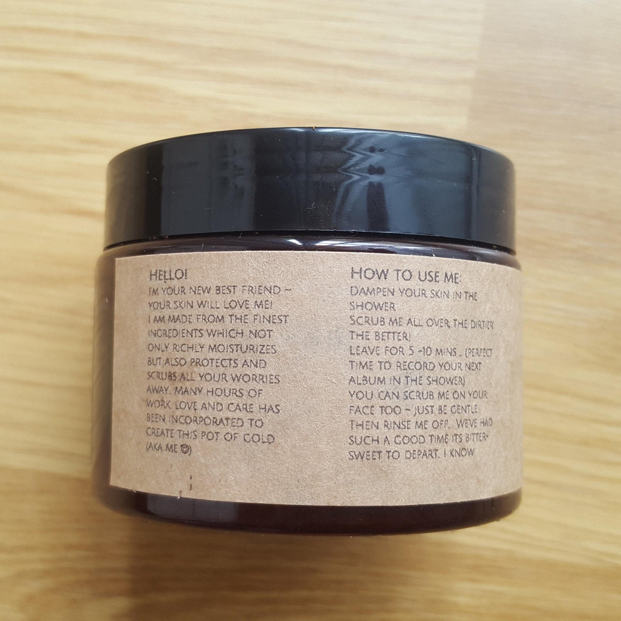 Body scrub jar with black lid, brown paper label, and text instructions on wood surface