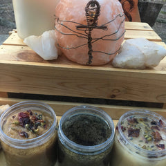 Body scrubs in clear jars with brown, green, and white contents, with dried flowers and crystals on wooden surface