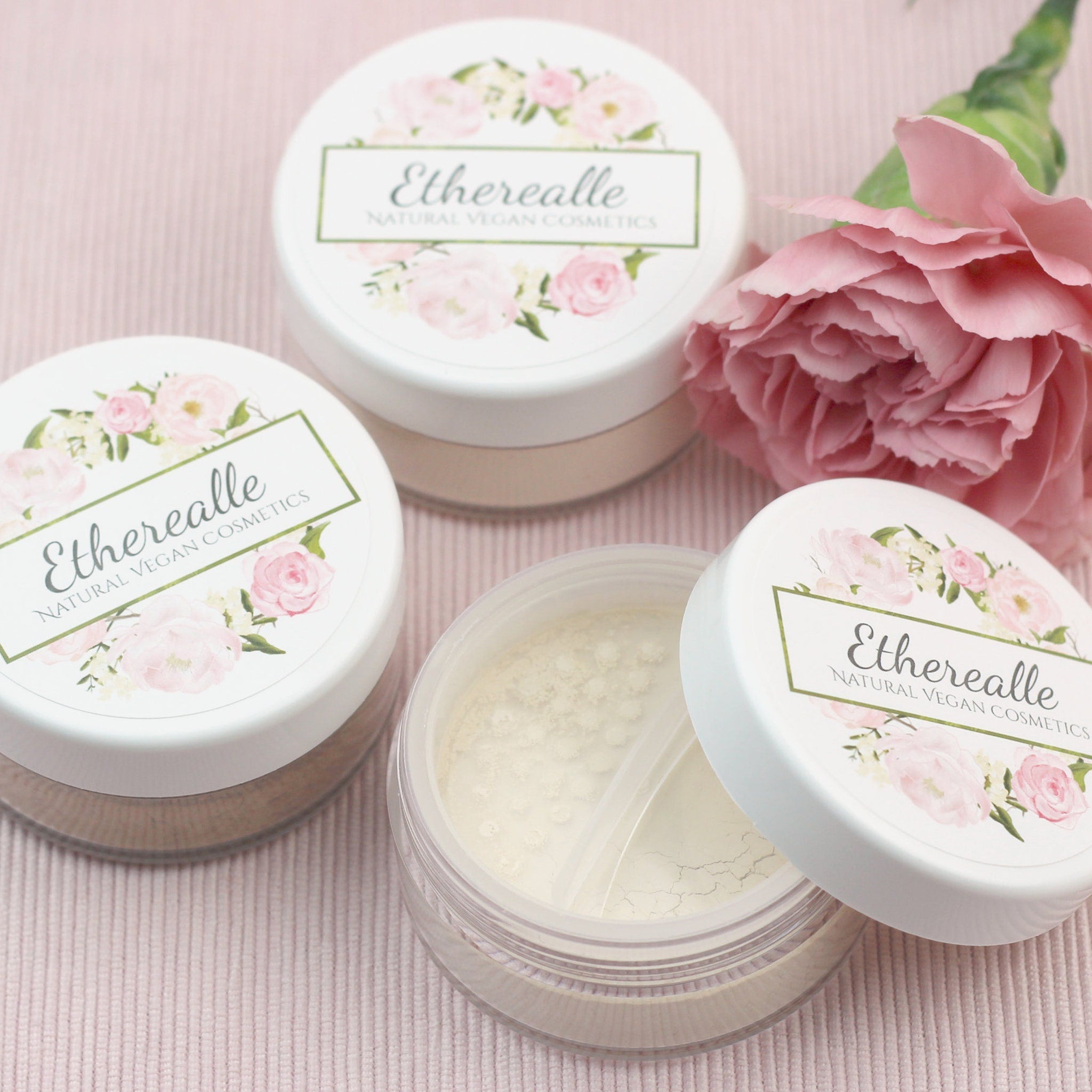 Cosmetic powder jars with white lids, floral design, and Etherealle brand name on pink background