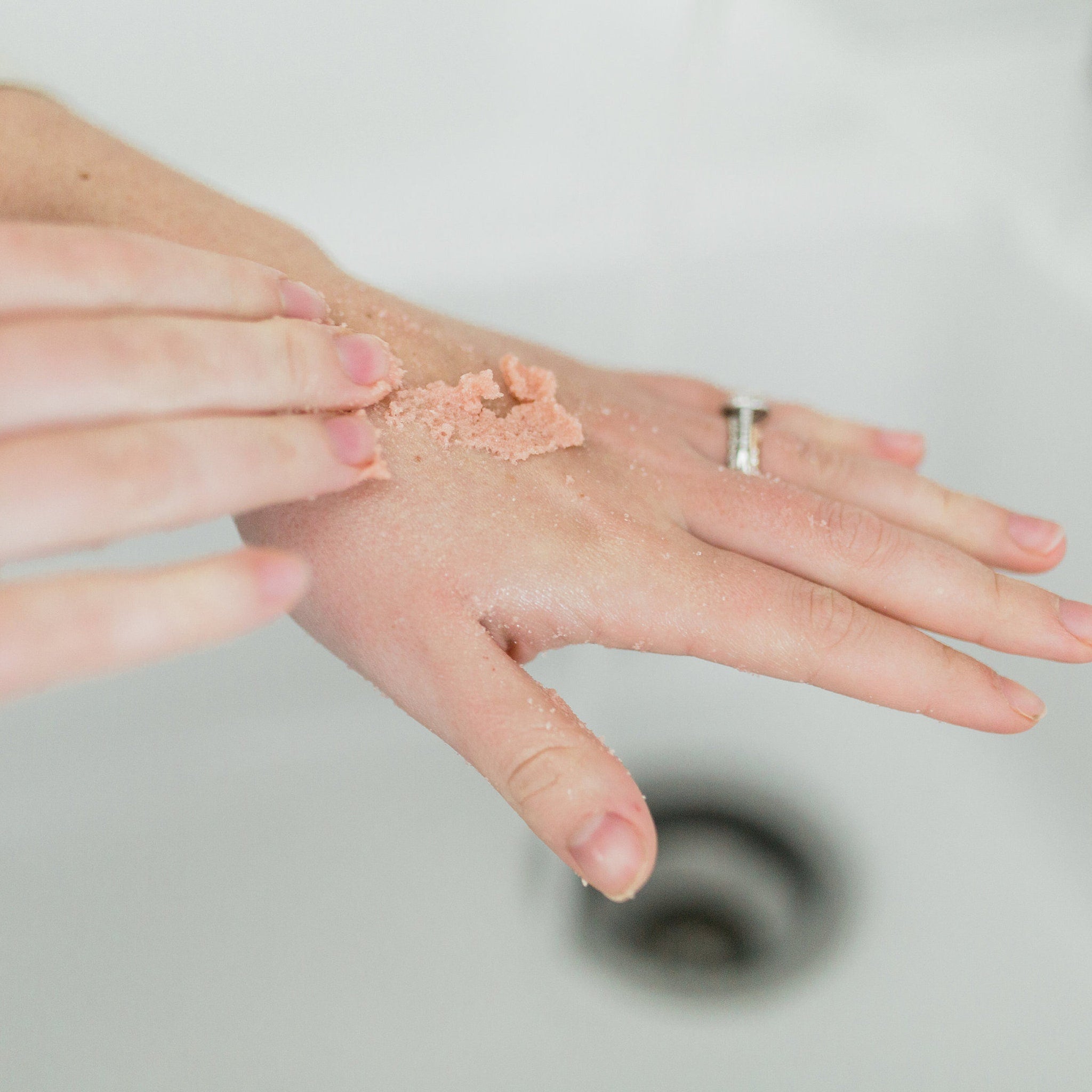 Hand scrub with pink granules and visible texture, being applied to skin over a white sink