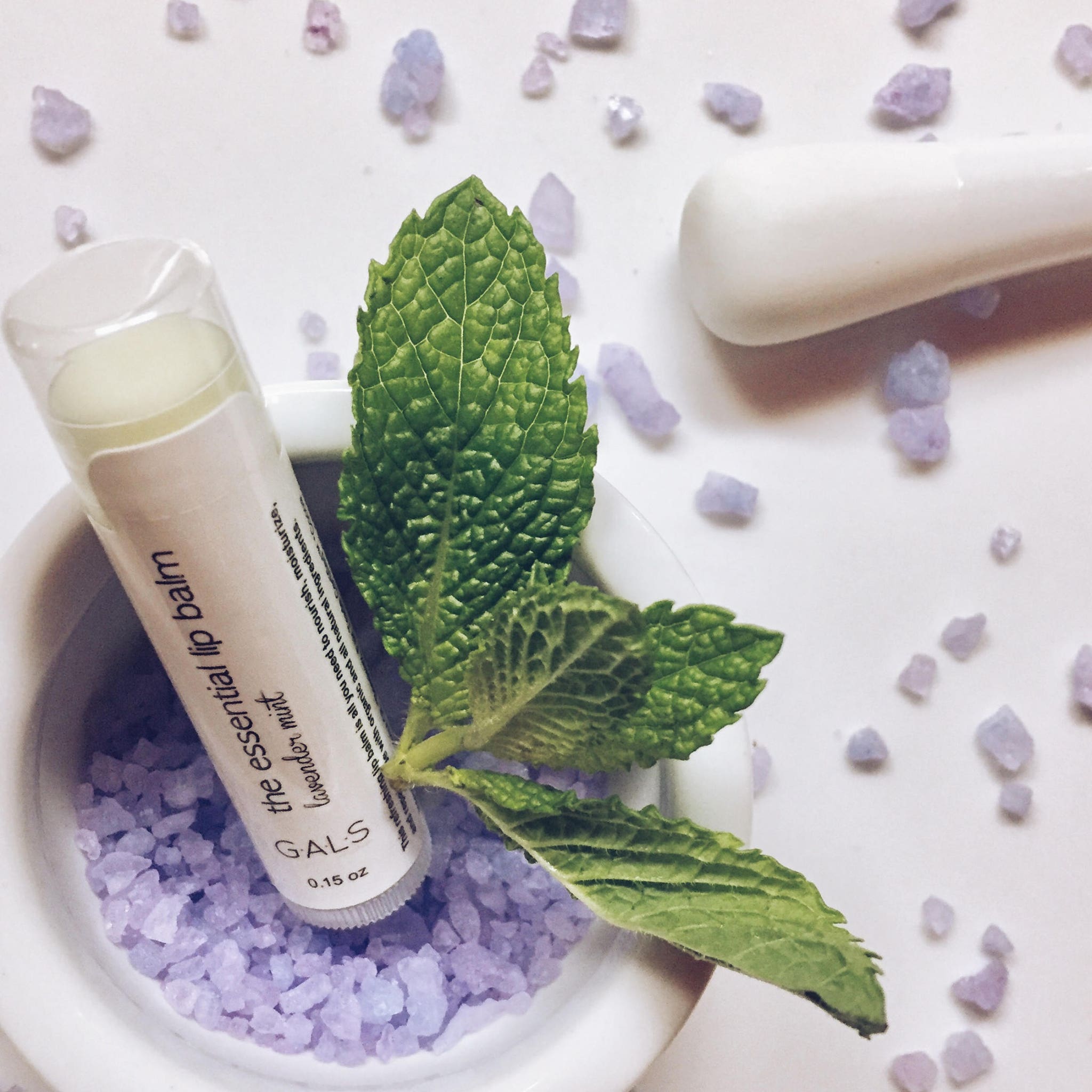 Lip balm tube with white label, mint leaf, and purple crystals in a white bowl