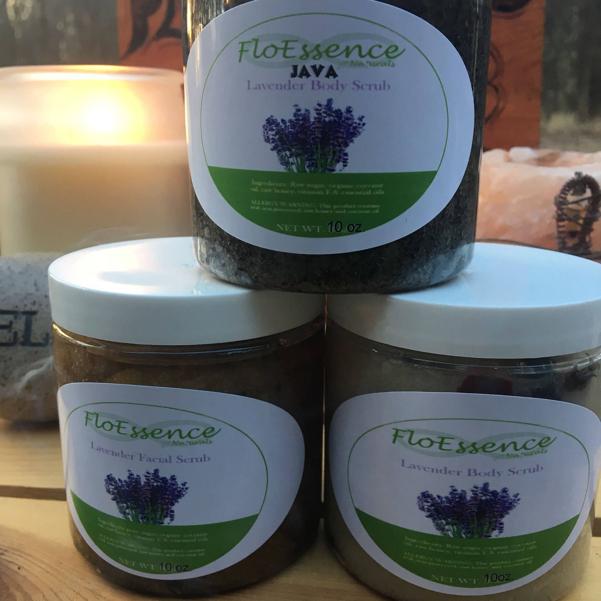 Body scrub jars with white lids, FloEssence labels, and dark textured contents, stacked on wood surface