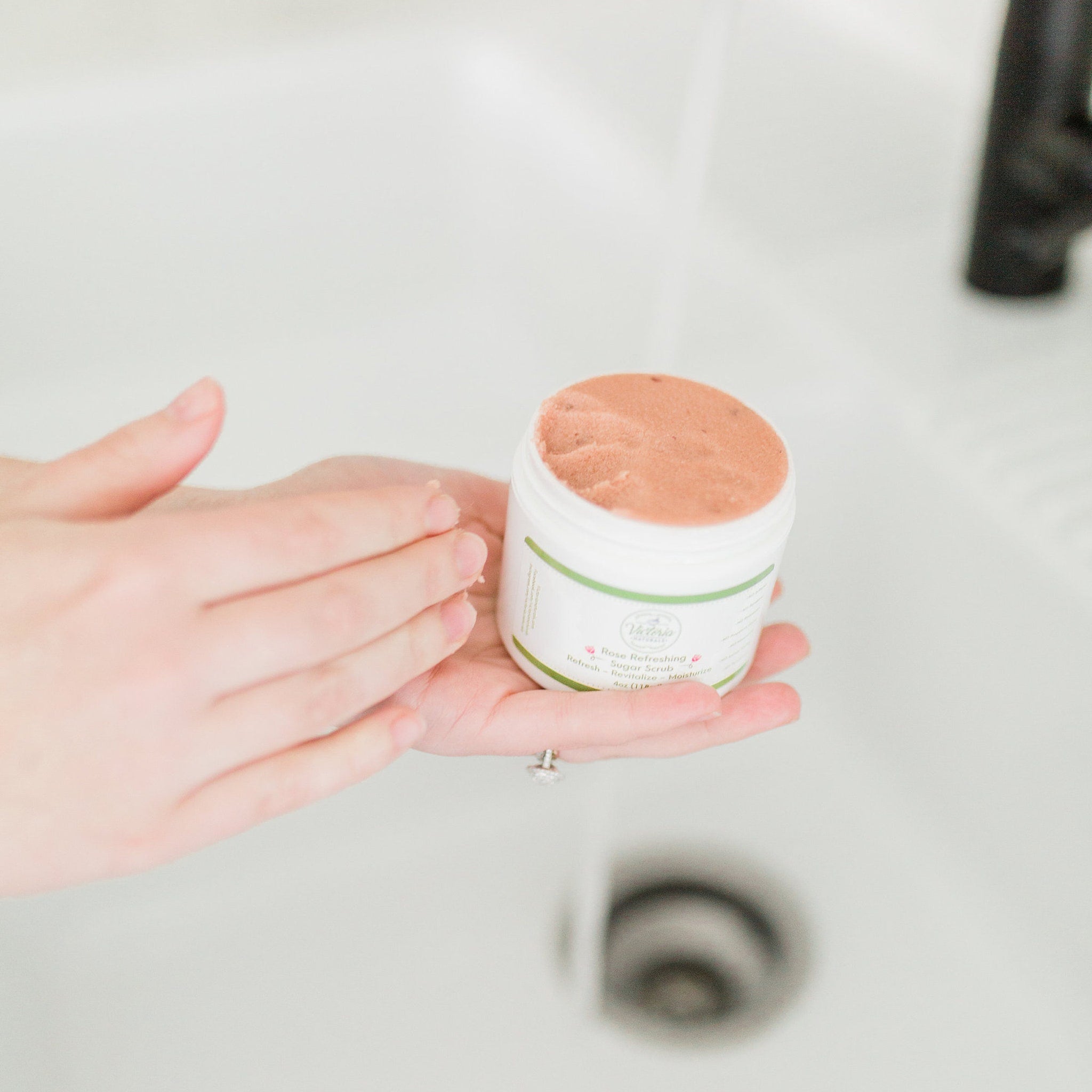 Sugar scrub jar with pink scrub, white jar, green label, held in hands under water