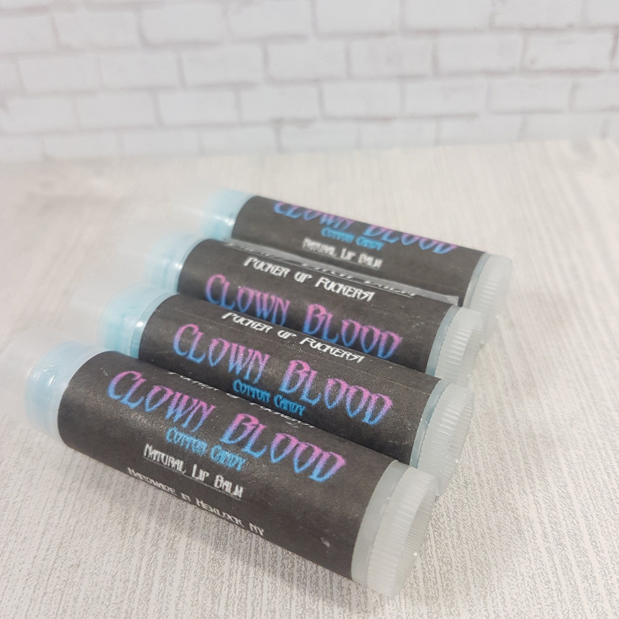 Lip balm tubes with black labels and blue/pink text, stacked on white surface, "CLOWN BLOOD" visible