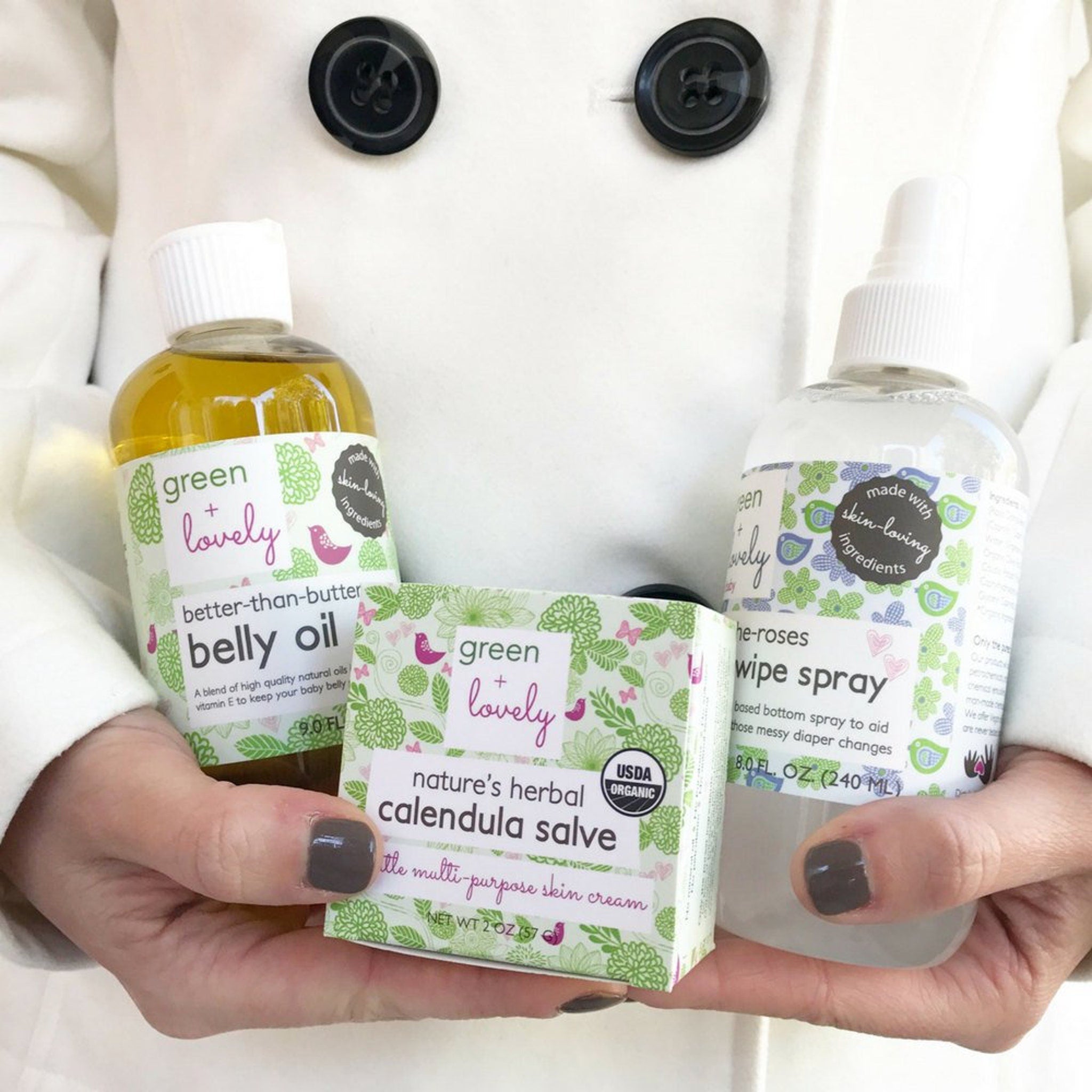 Baby skincare products with green and white labels, held in hands with dark nail polish, and white coat background