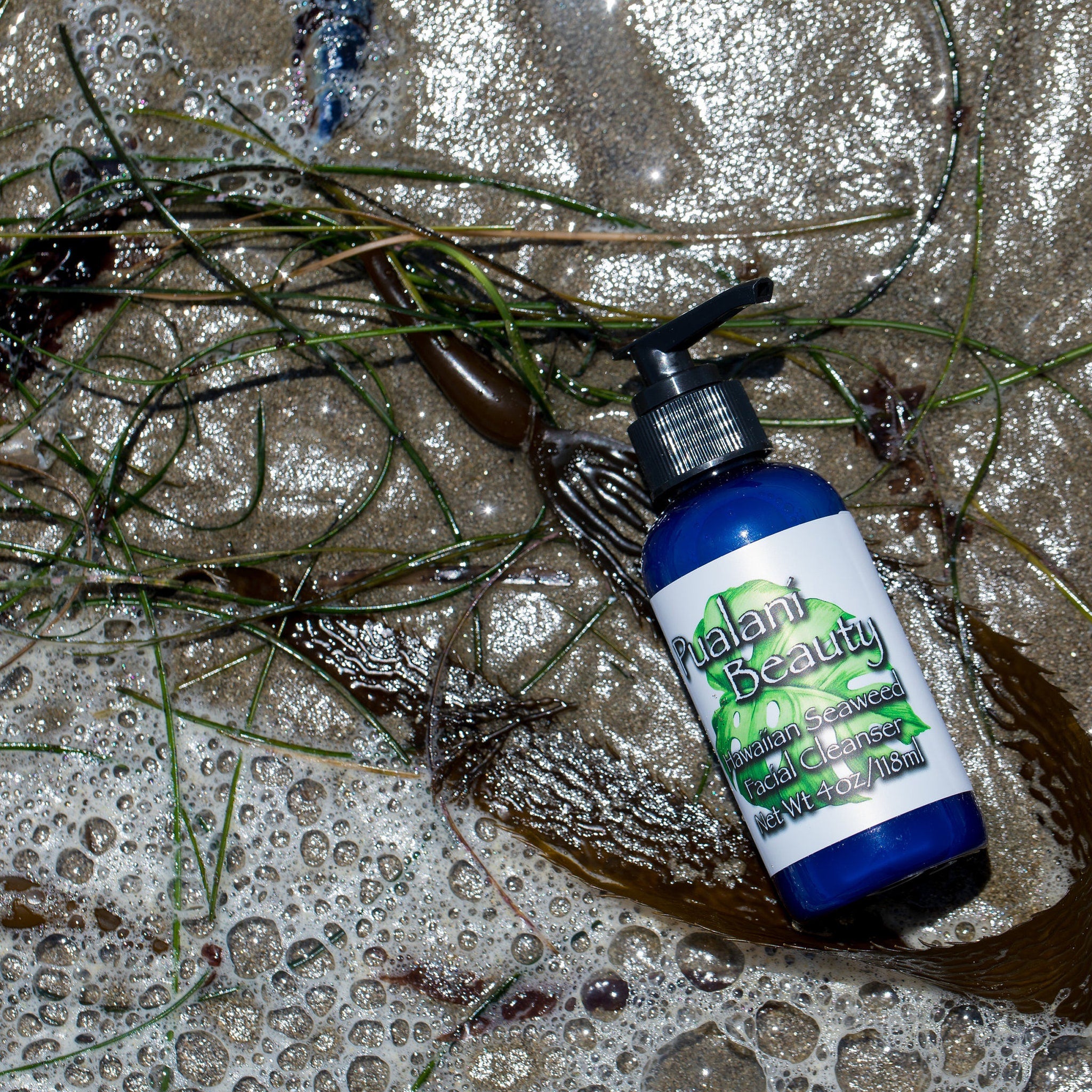Facial cleanser bottle, blue and white, with "Pualani Beauty" label, on sand with seaweed