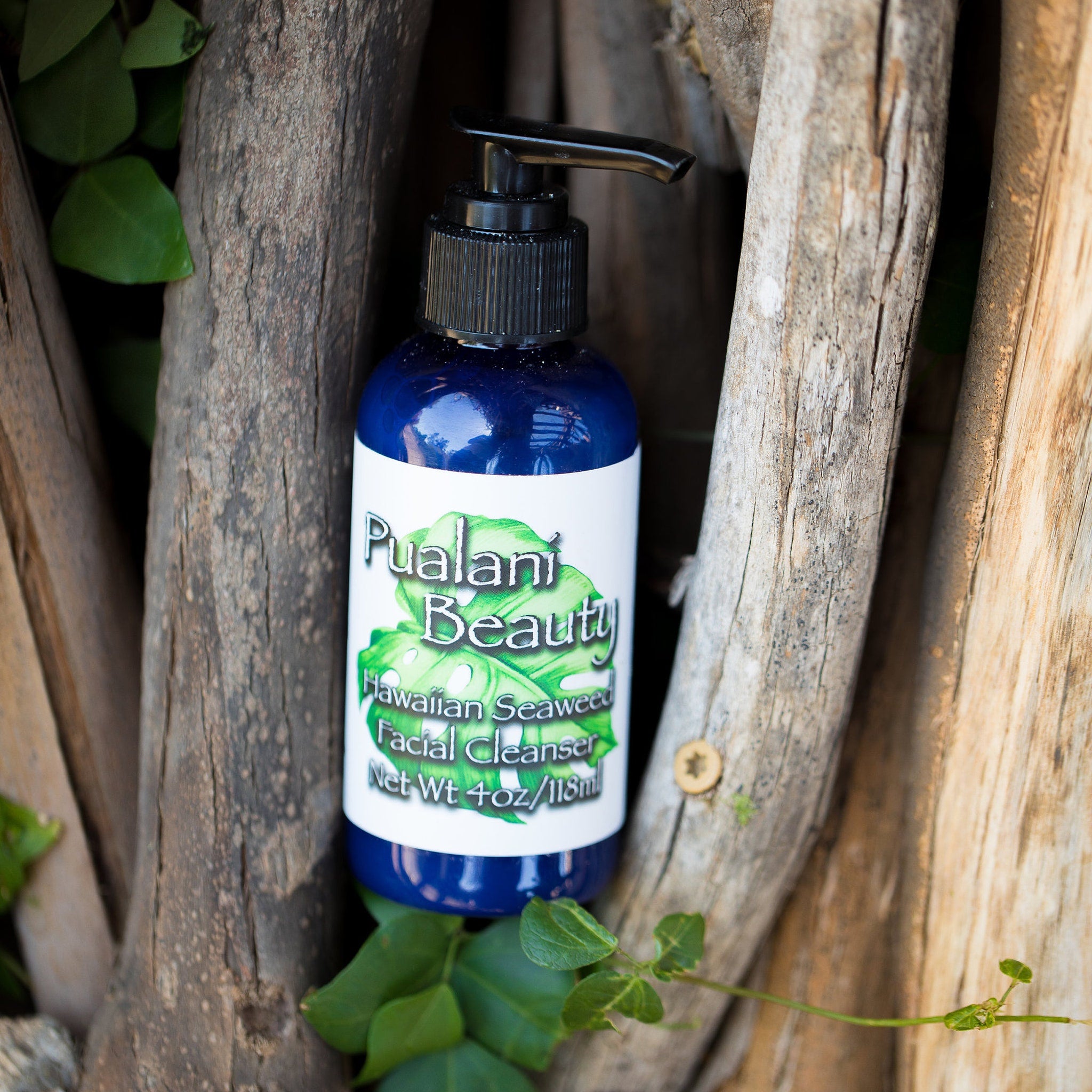 Facial cleanser bottle, blue glass, black pump, Pualani Beauty label, set against wood branches