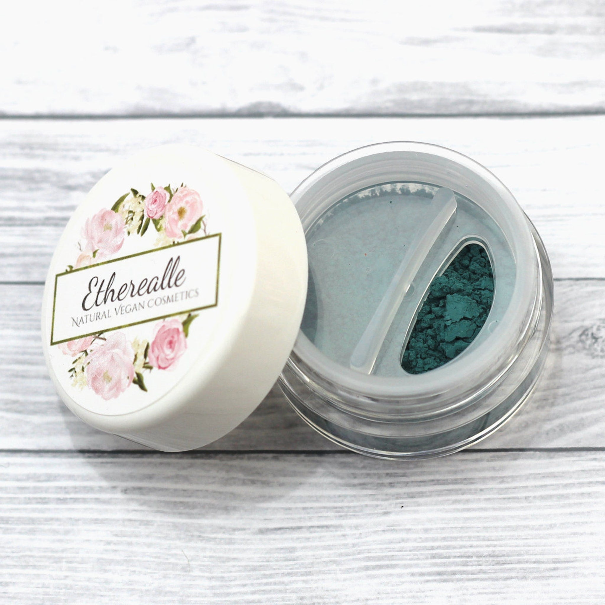 Eye shadow jar with white lid, floral design, and teal powder inside clear container