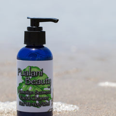 Facial cleanser bottle, blue glass, black pump, Pualani Beauty label, on beach