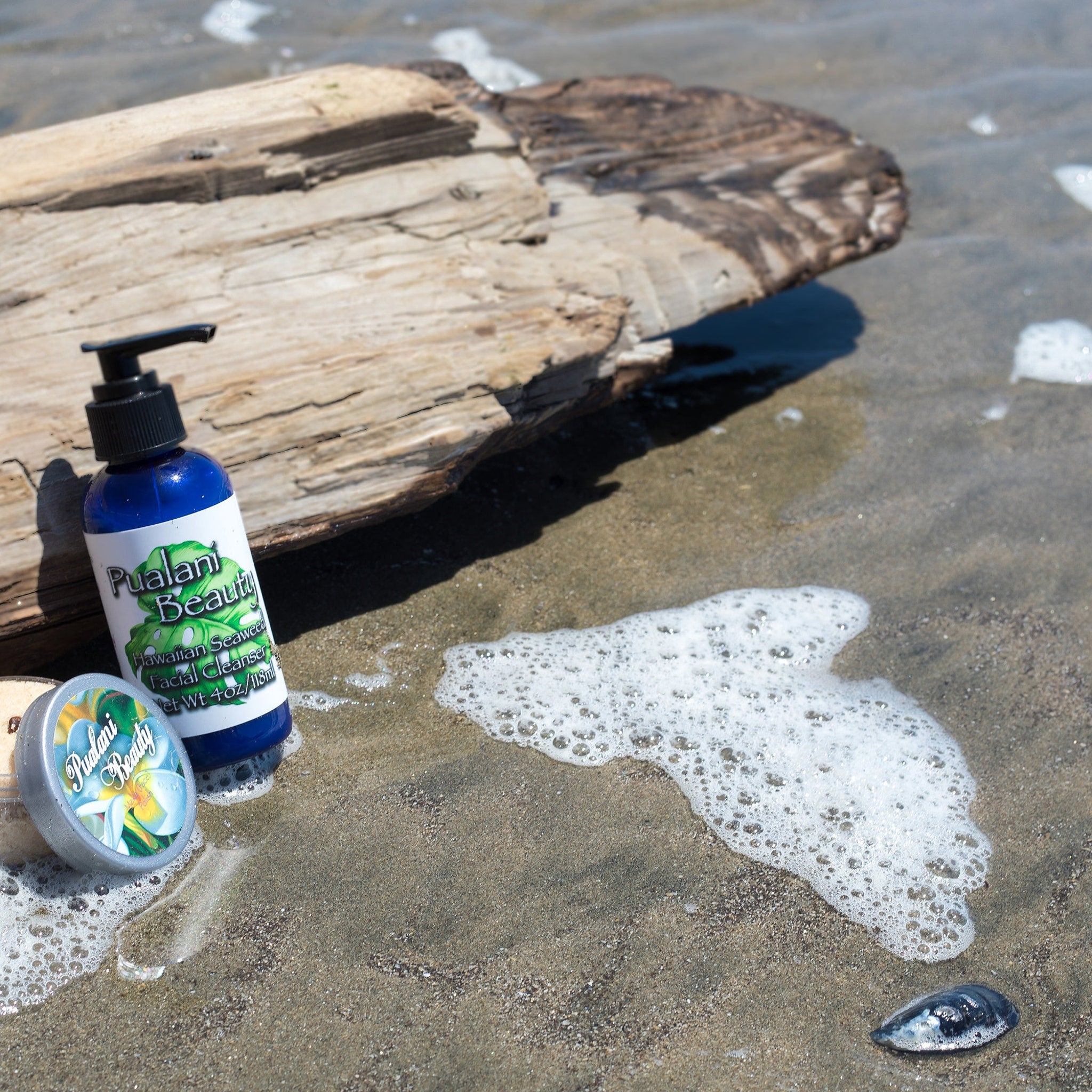 Pualani Beauty blue bottle with black pump, silver lid, on sandy beach with driftwood