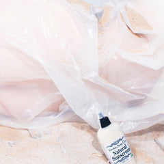 Natural sunscreen bottle with white and black cap, sand, and clear plastic bags, Veritas Soap Co label