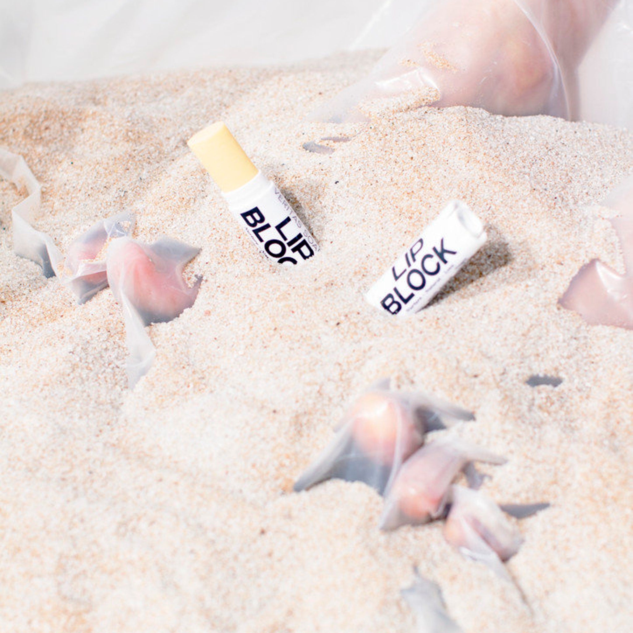 Lip block tubes with white and yellow accents, "LIP BLOCK" text on sand with plastic wrap