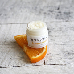 Lip butter jar with white cream, orange slice, and Bellaroma branding on white wooden surface