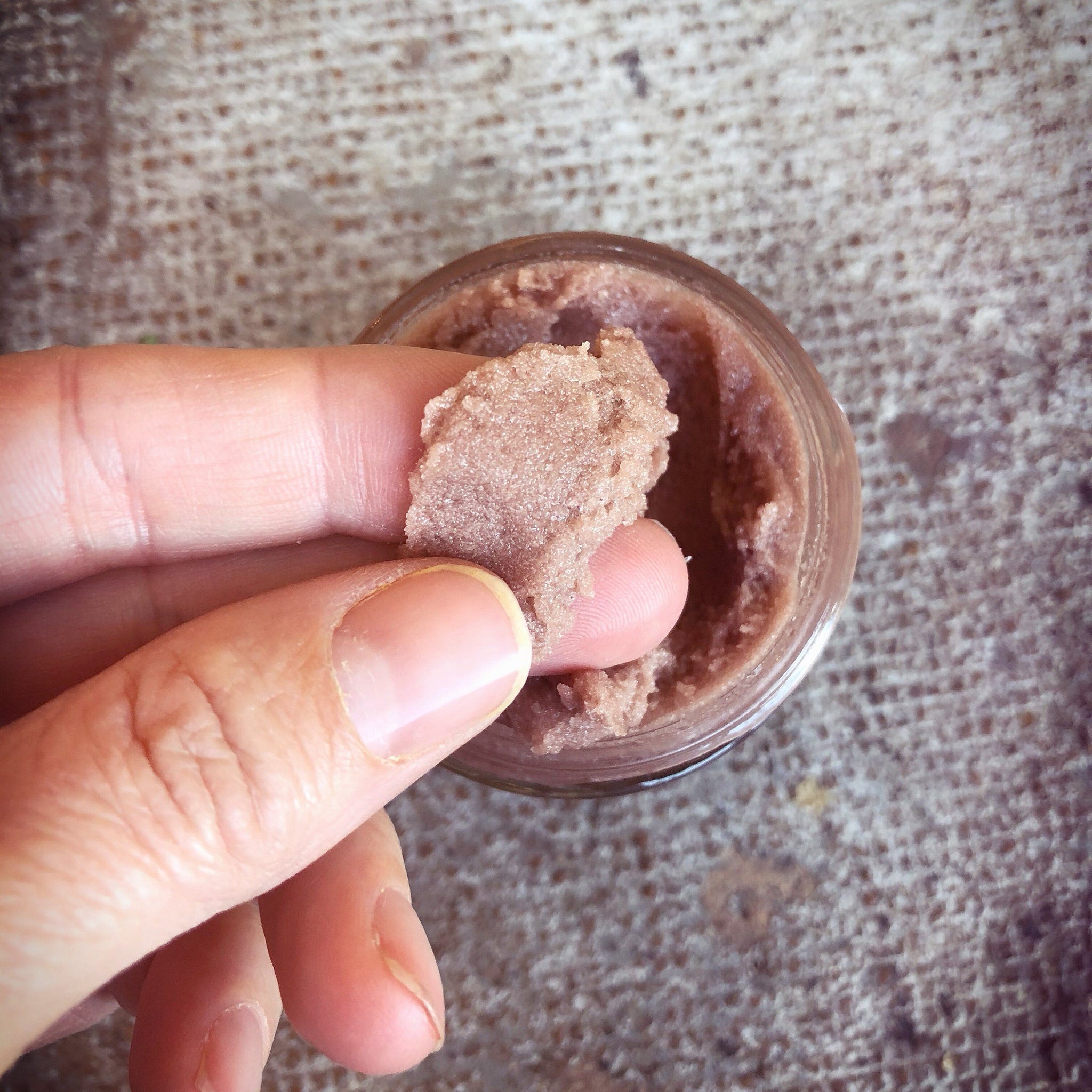 Exfoliating scrub with pinkish brown color, grainy texture, held by hand, in jar