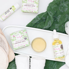 Calendula skin care products in green and white packaging, with green fabric, and a serum bottle with a dropper.