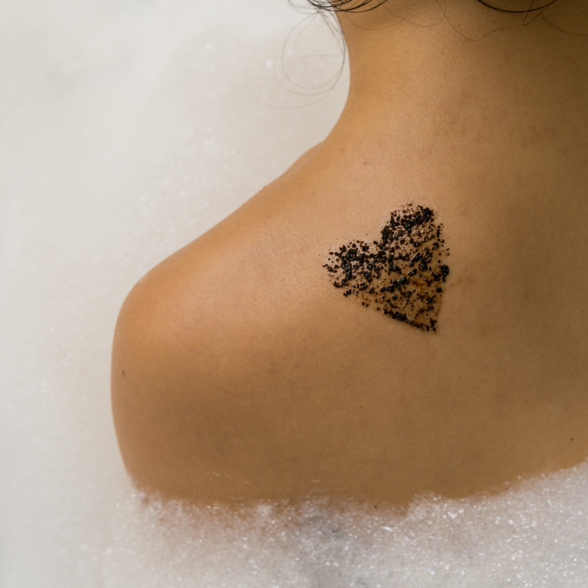 Body scrub application on shoulder with heart-shaped pattern in bath bubbles