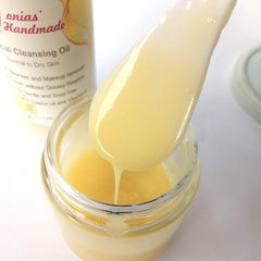 Facial cleansing oil, pale yellow liquid, pouring from a spoon into a glass jar, "onias' Handmade" visible