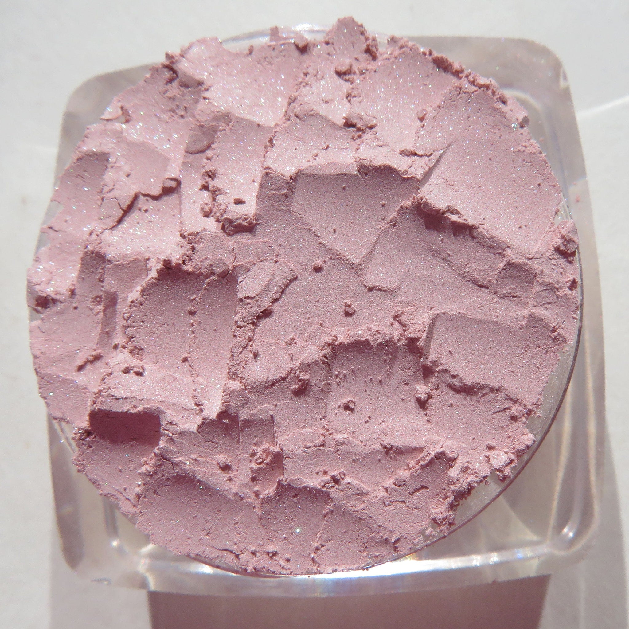Side view of pastel pink loose powder eyeshadow in a transparent jar with black screw-top lid, showcasing fine shimmer particles.
