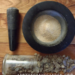 Mortar and pestle set with black stone, beige powder, and ApothecarysGarden text on wood surface