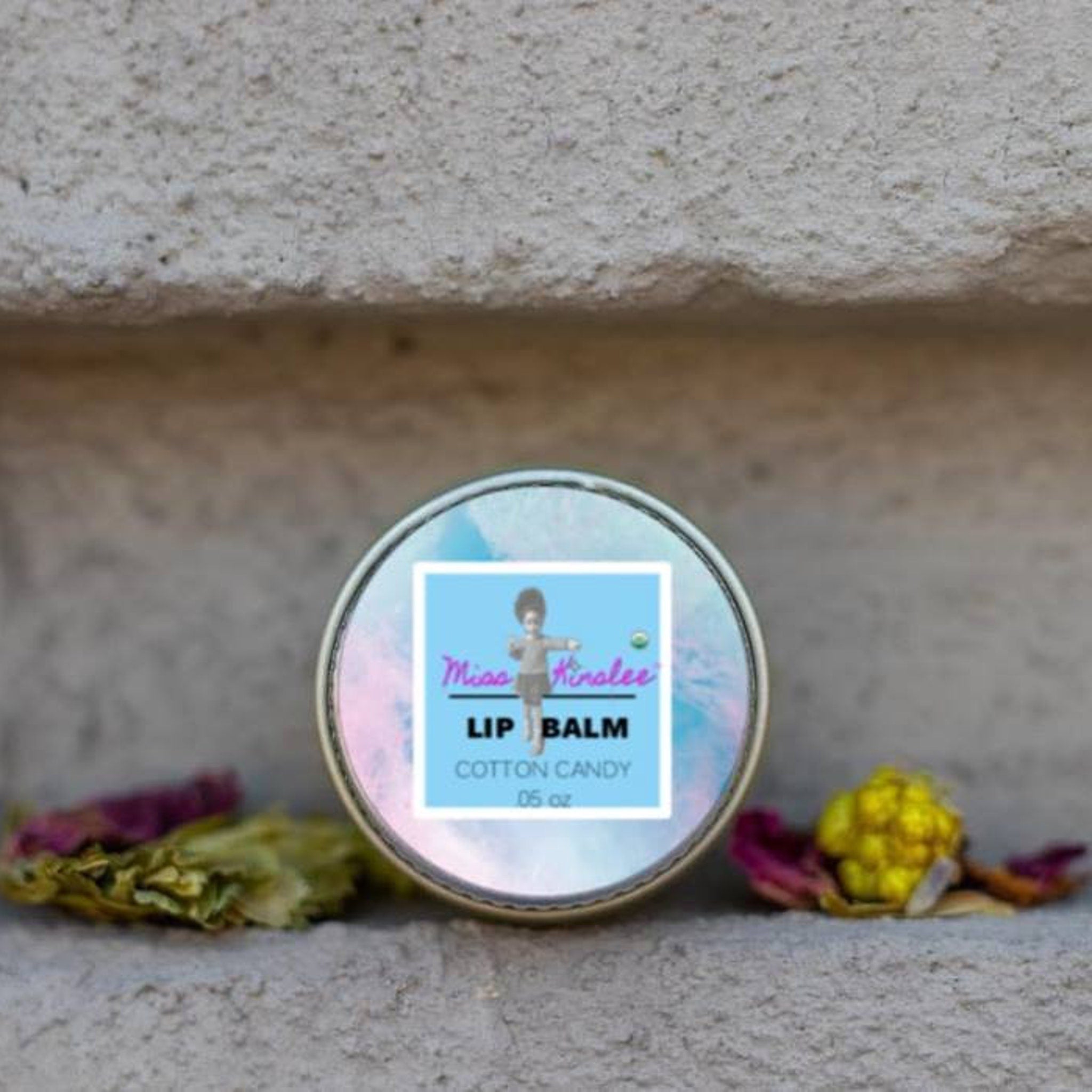 Lip balm tin with iridescent lid, blue label, and Miss Kirolee text, on a textured surface