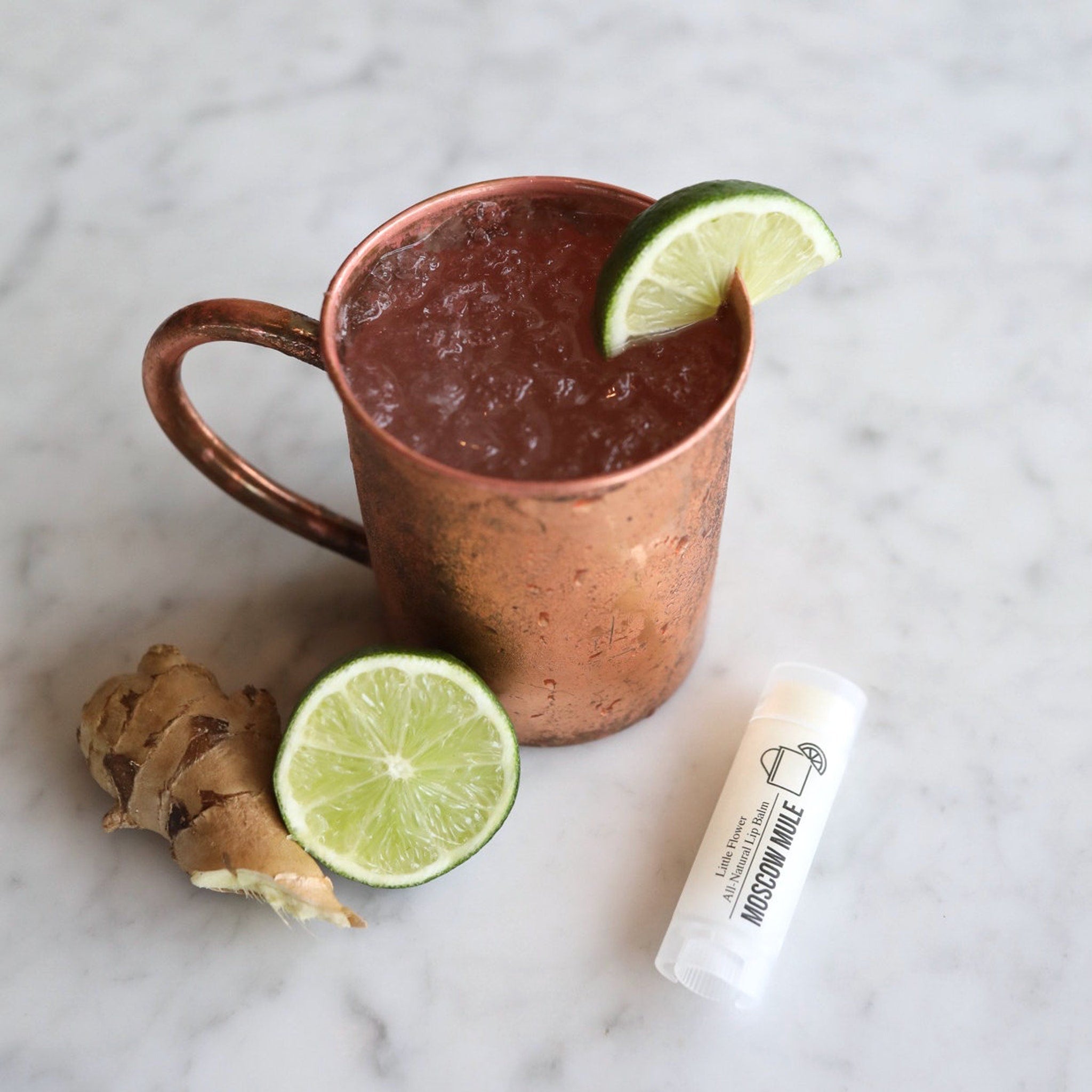Moscow Mule lip balm with white tube, text, and lime slice garnish on marble surface