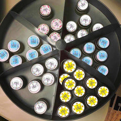 Lip balm tins with various label designs, round metal containers, displayed in a divided tray.