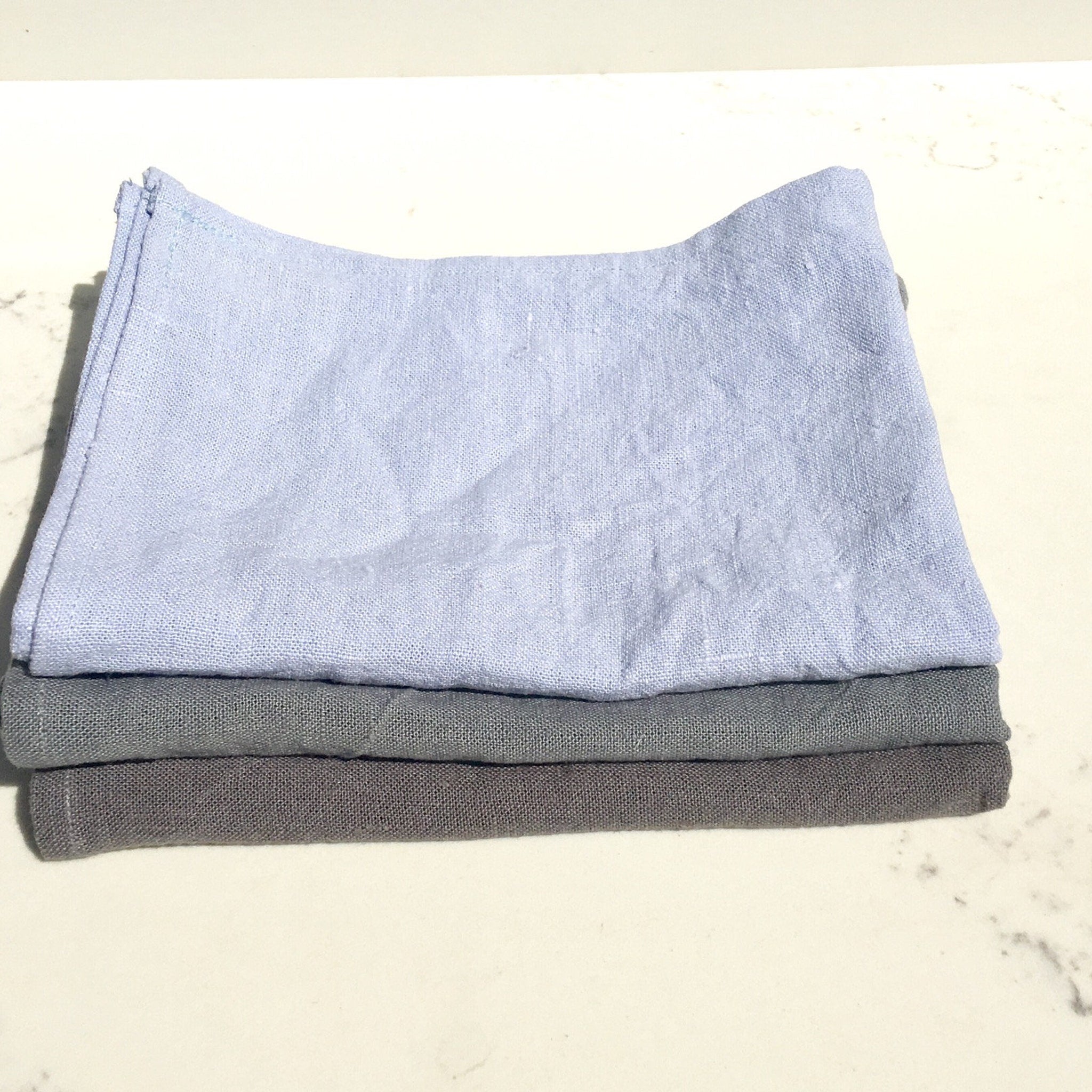 Linen cloths in light blue, gray, and dark gray, stacked on top of each other