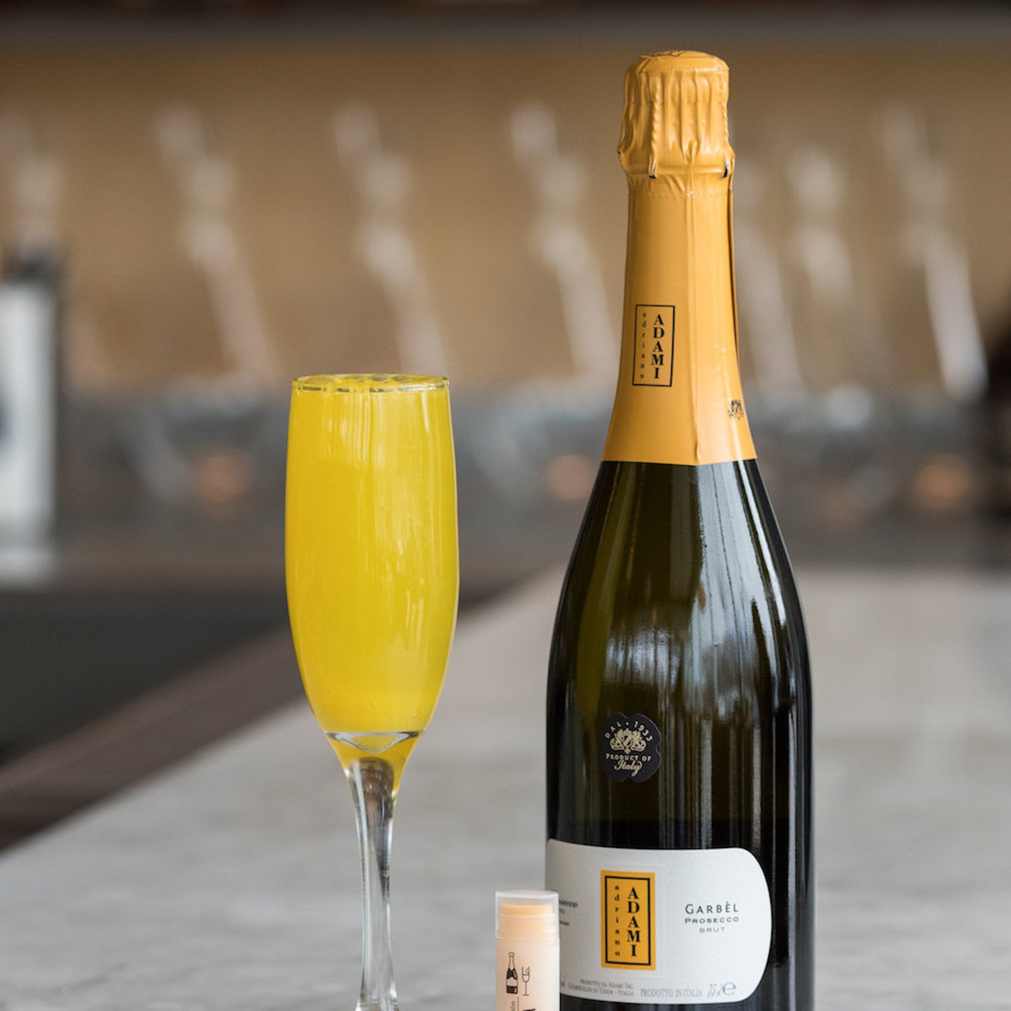Mimosa cocktail in tall glass with yellow liquid, Adamì Prosecco bottle, and lip balm on counter