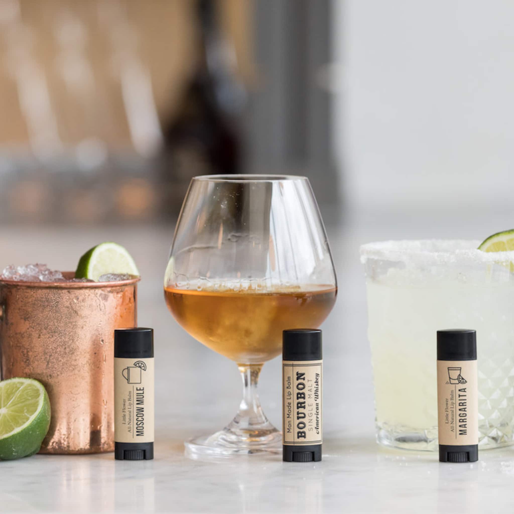 Lip balm sticks with black caps, tan labels, and cocktail names near drinks