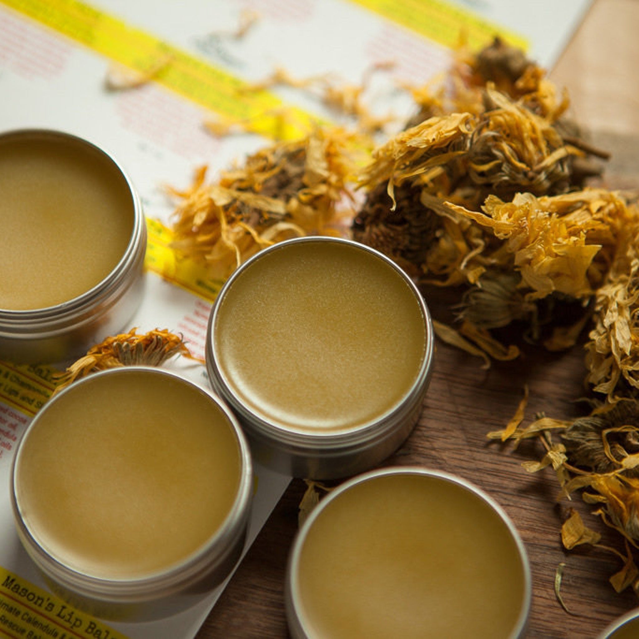 Lip balm tins with yellow balm, silver metal containers, and dried flowers on wood surface