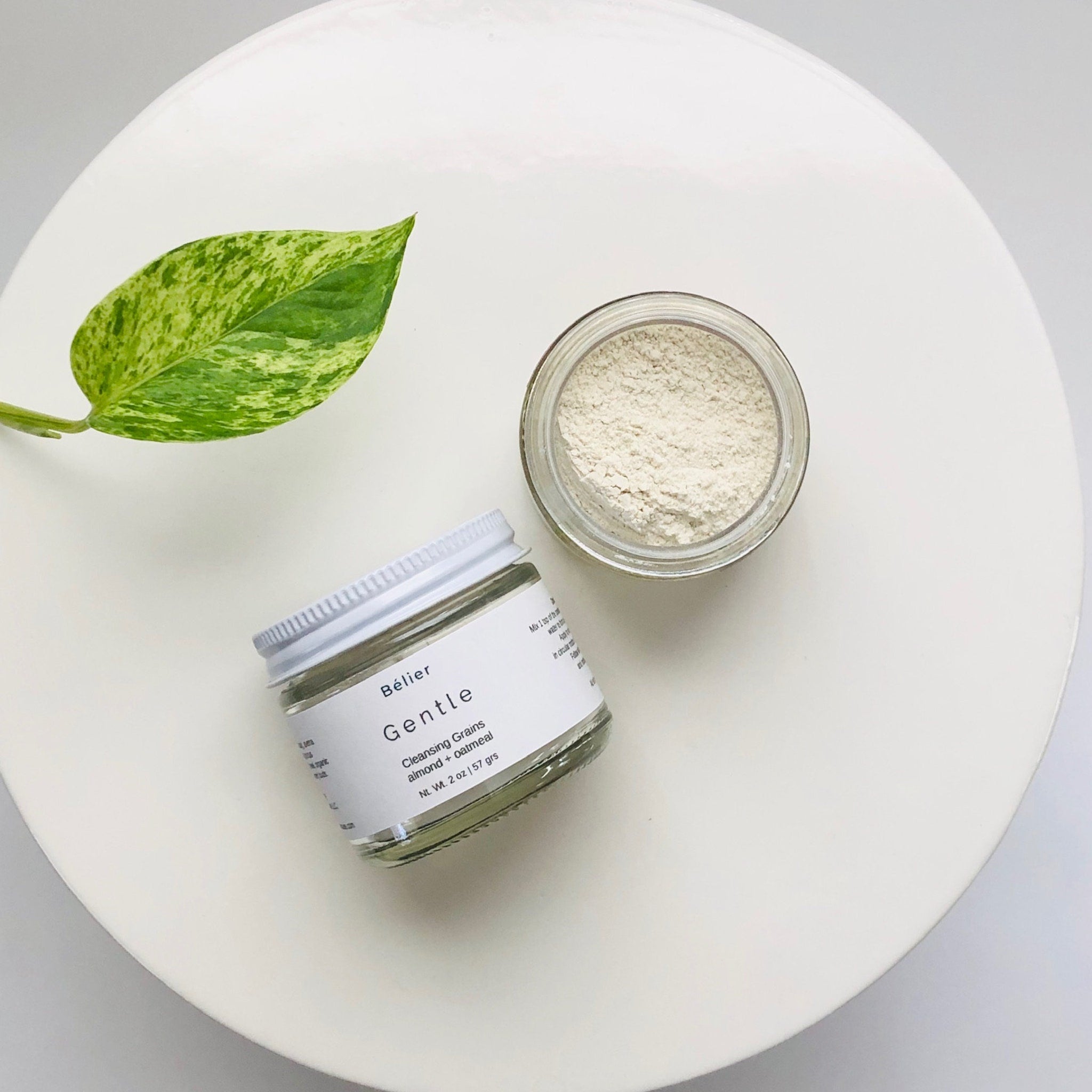 Facial Cleanser in glass jar with white lid, beige powder, and green leaf on white surface