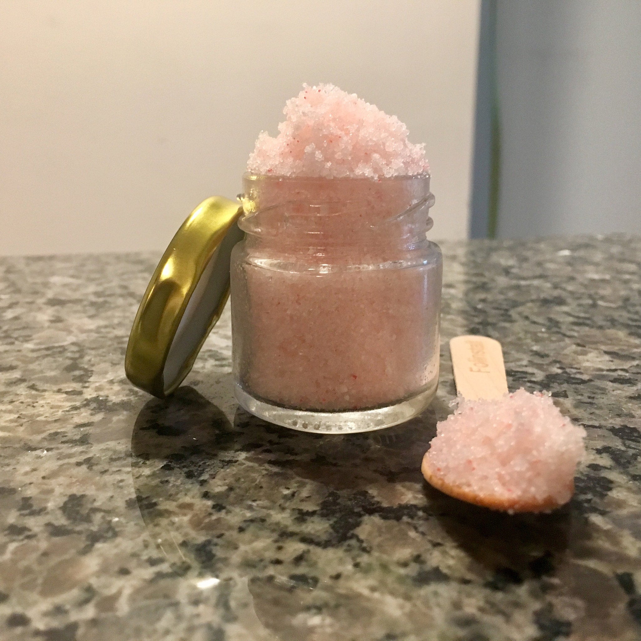 Pink scrub in glass jar with gold lid, wooden scoop with scrub on counter