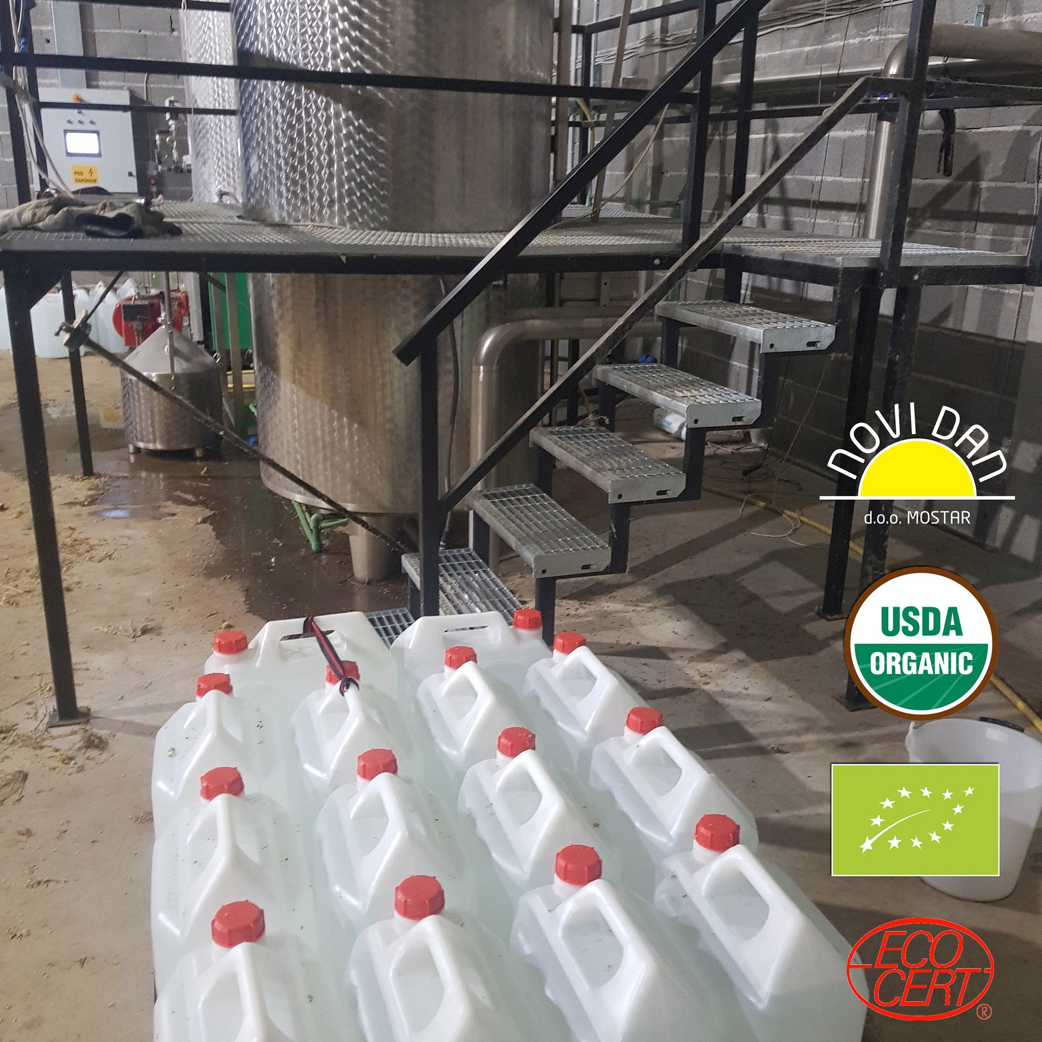 White plastic containers with red caps, stacked on concrete floor near metal stairs and tanks, New Day logo visible
