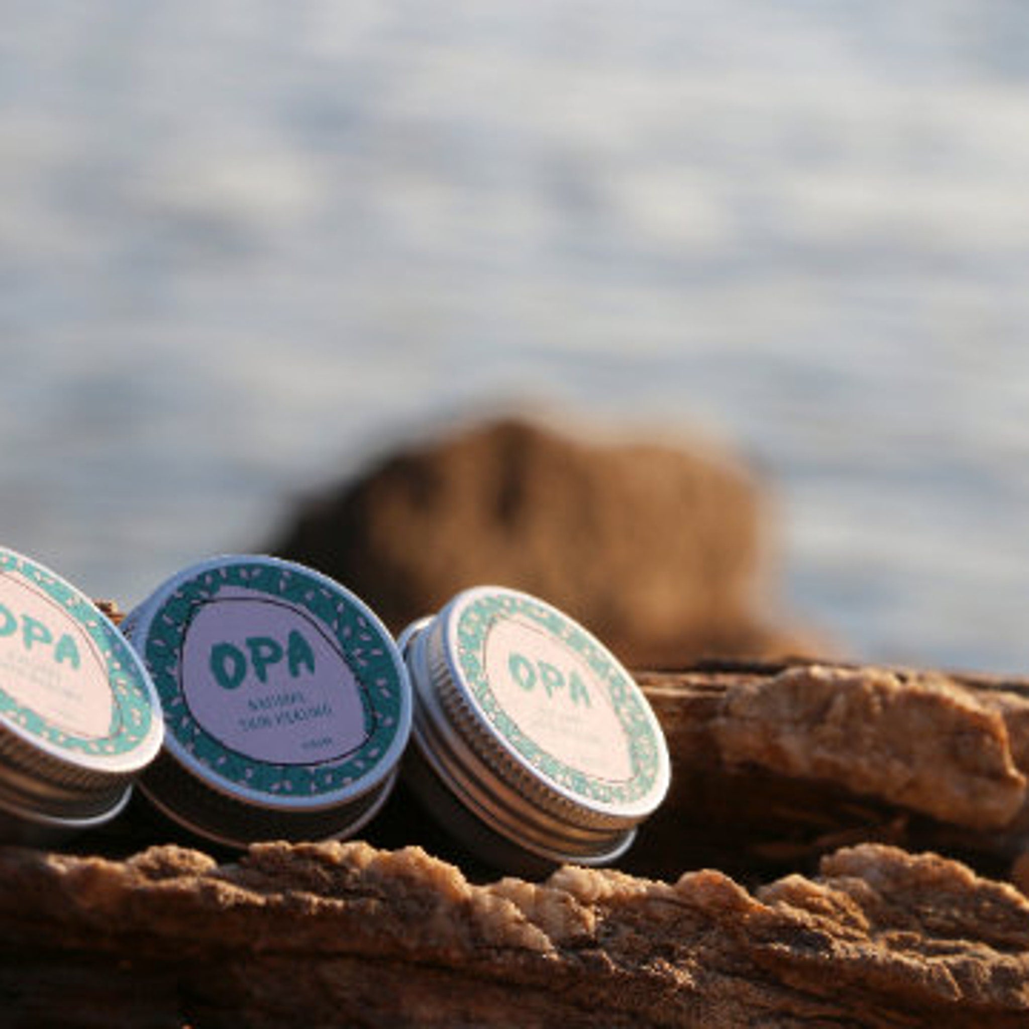 OPA skincare tins with teal and silver lids on a wooden surface near water