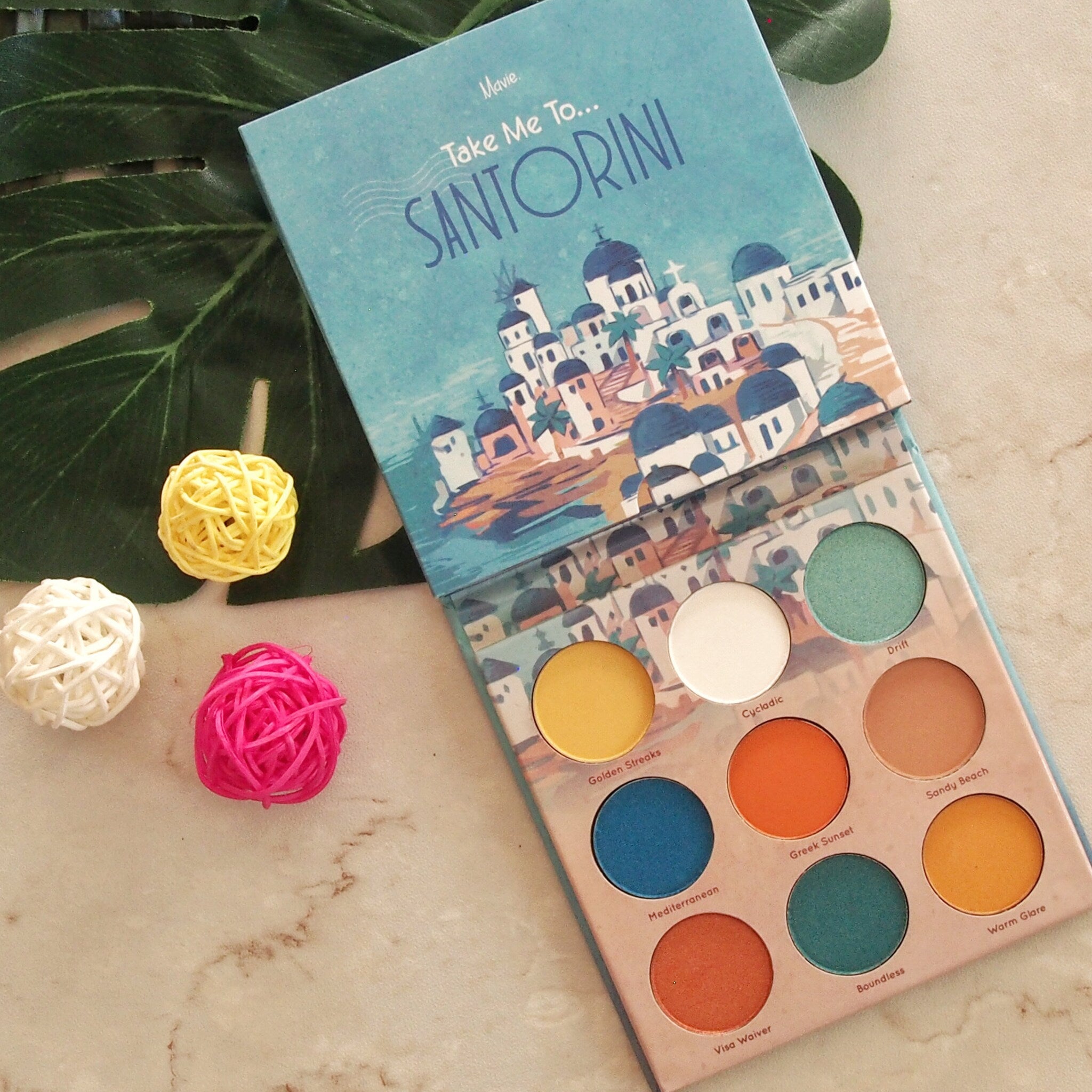 Eyeshadow palette with blue, yellow, and orange shades, printed "Take Me To...SANTORINI" on the cover, and decorative balls