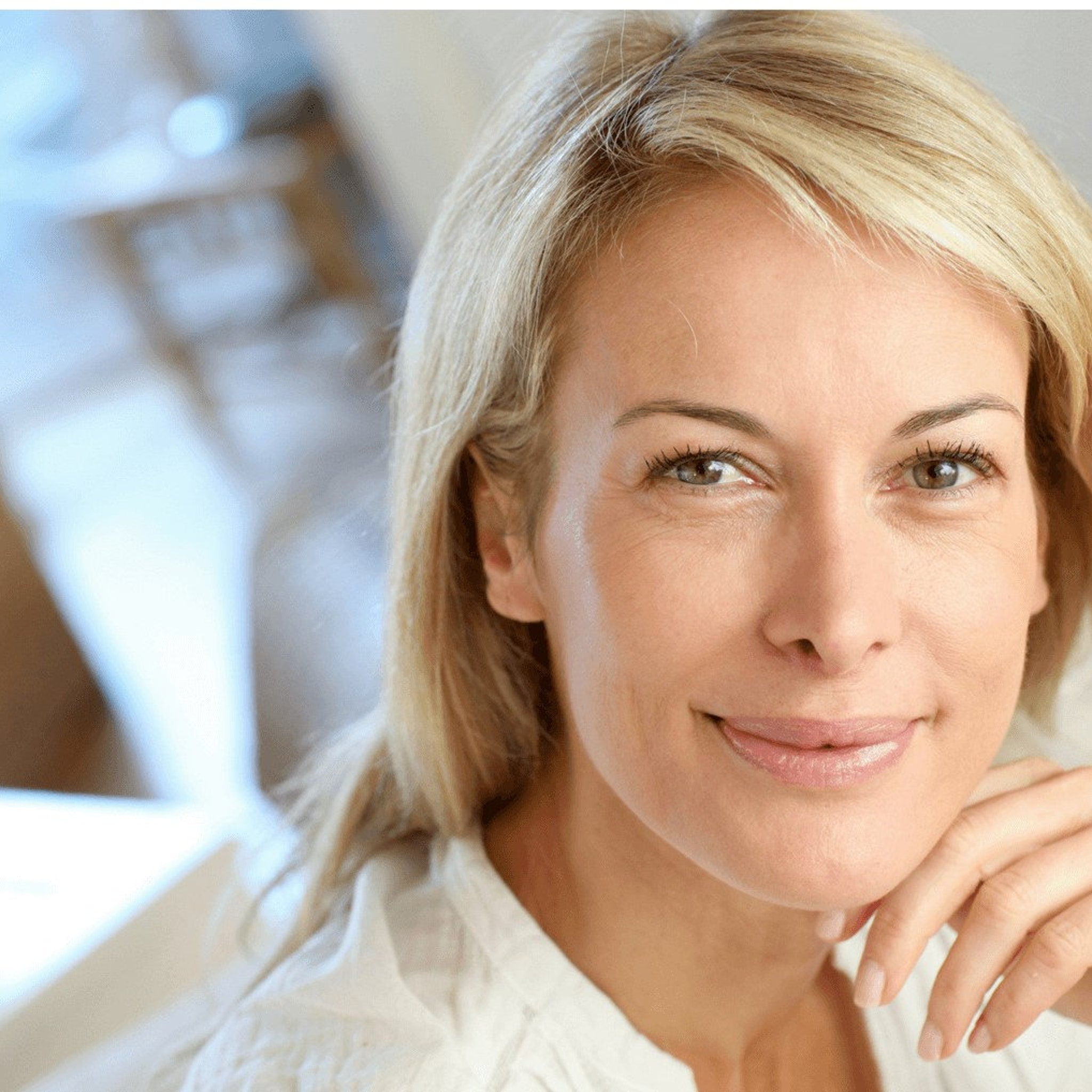 Woman with blonde hair and white shirt smiling, hand on chin, looking at camera