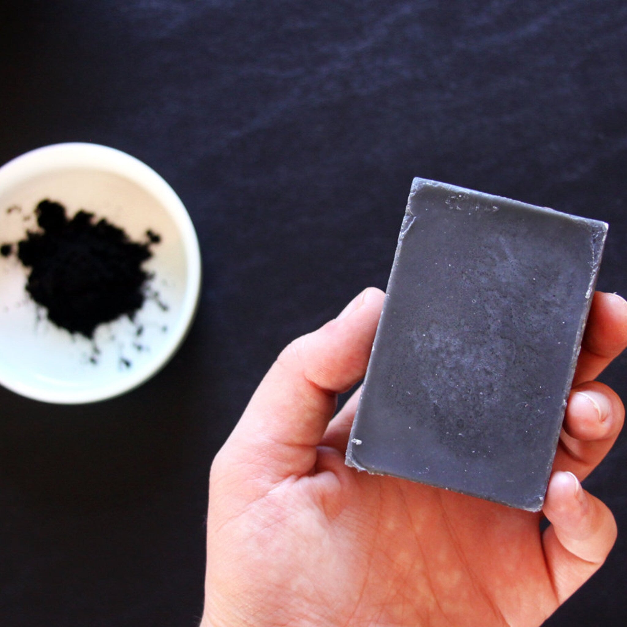 Soap bar held in hand, gray rectangular shape, with bowl of black powder in background