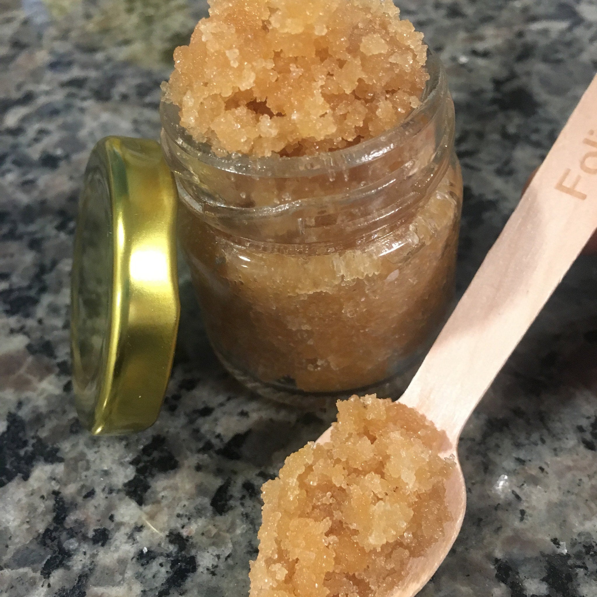 Sugar scrub in a glass jar with a wooden spoon, light brown granules, gold lid, and wooden spoon