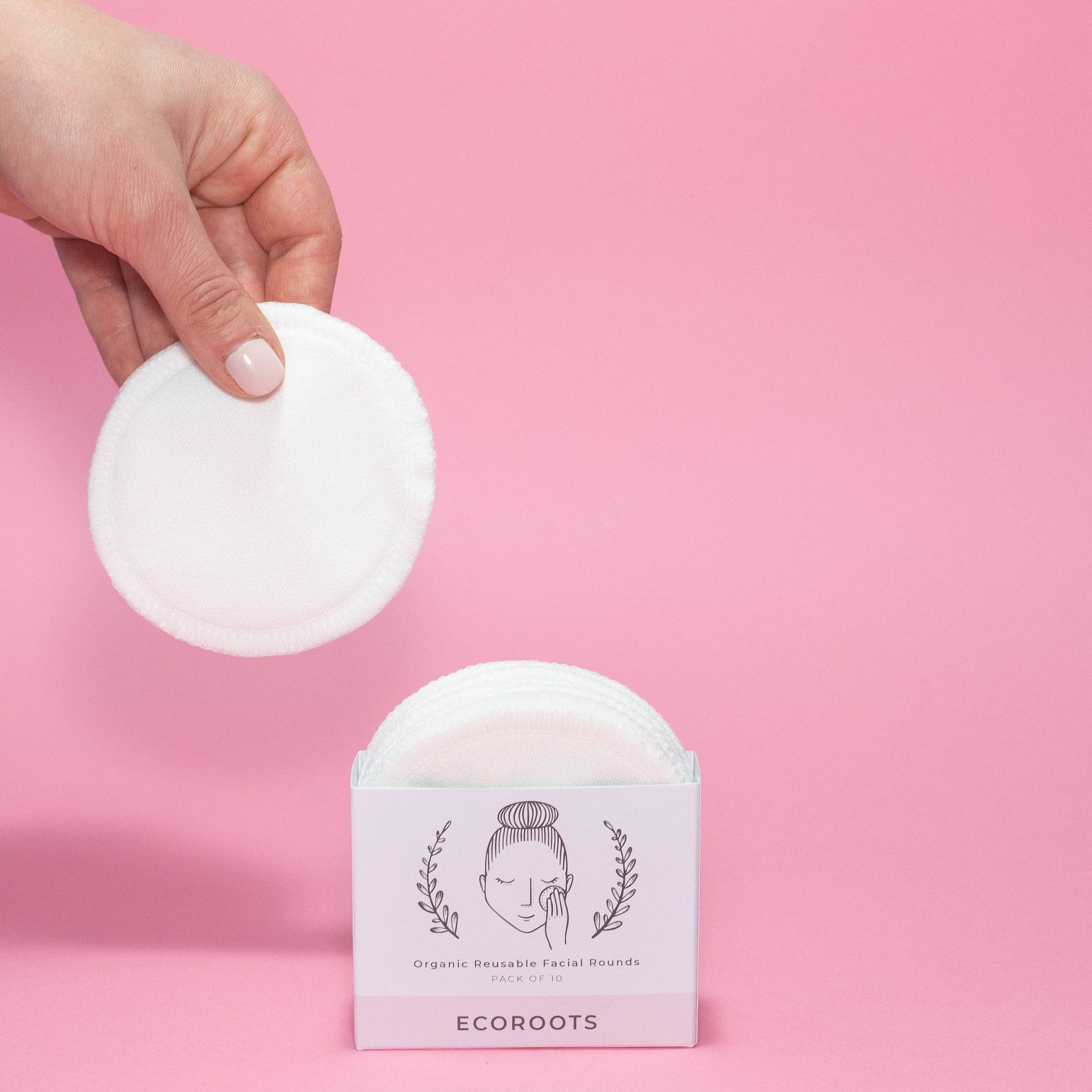 Reusable facial rounds, white cotton pads, held by hand, with Ecoroots box on pink background