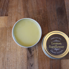 Lip balm in metal tin with yellow balm, Herbs and Soil Lemon Sunflower label, on wood surface