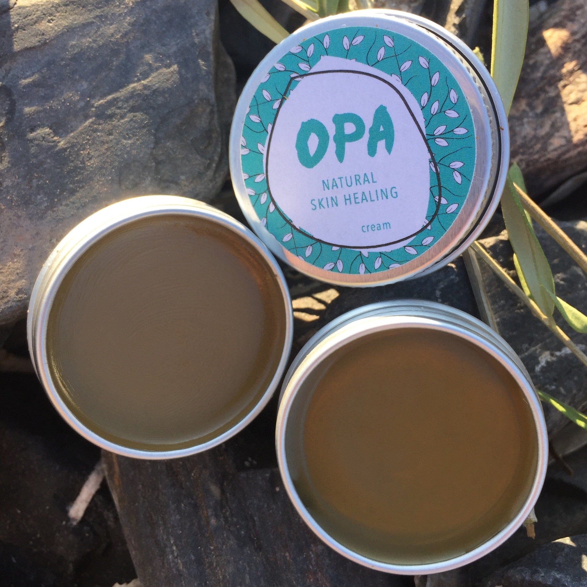 Skin cream in silver metal tins with OPA branding and green lid design, on rocks