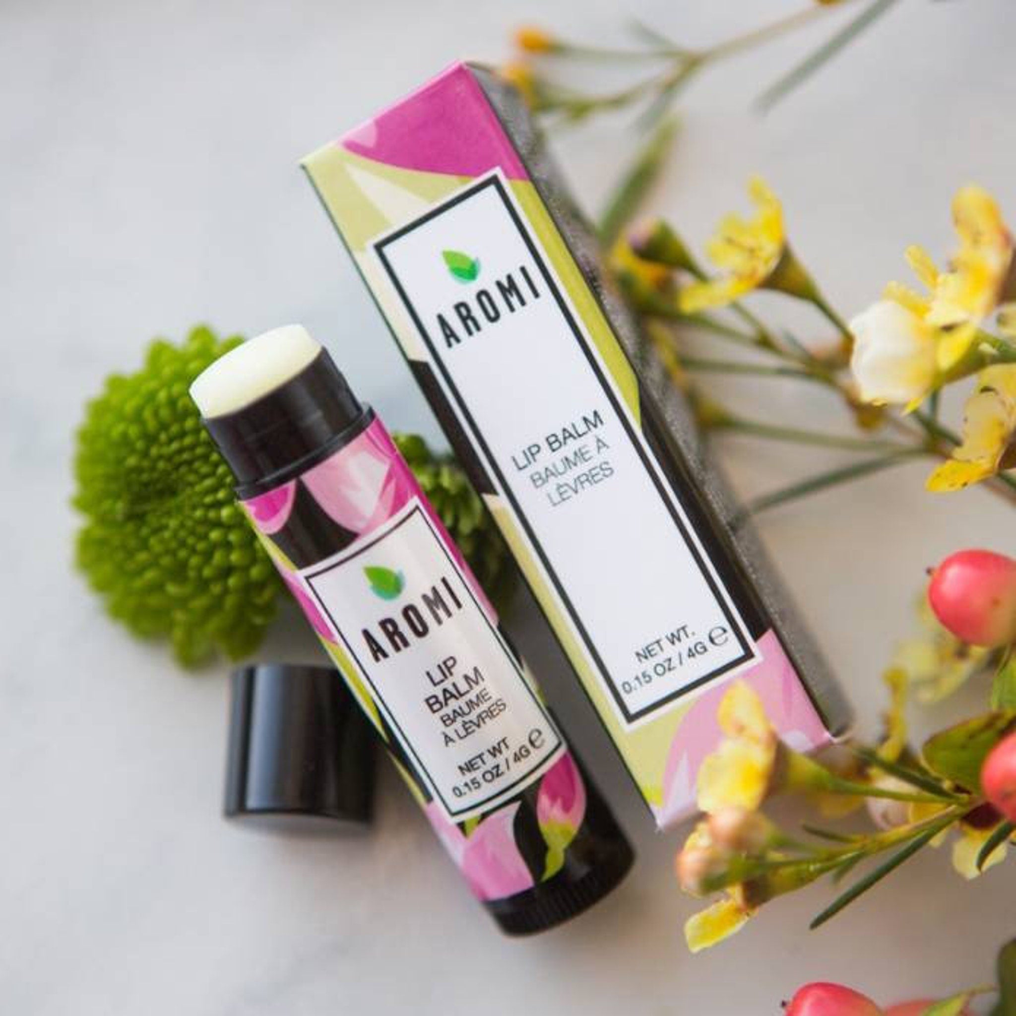 Lip balm tube with floral pattern, black cap, and AROMI brand text, with box and flowers