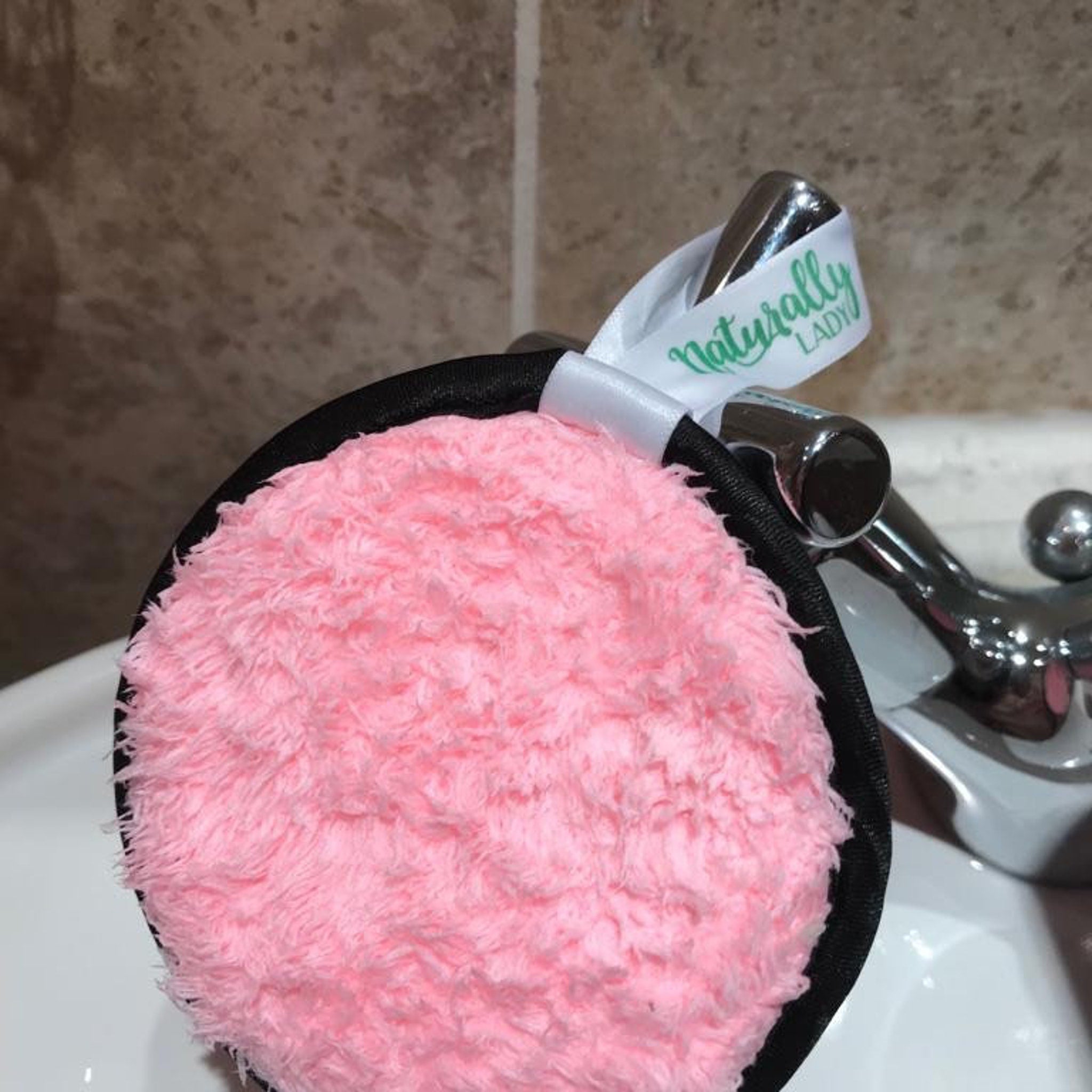 Makeup remover pad with pink fluffy material, black trim, and Naturally Lady ribbon detail